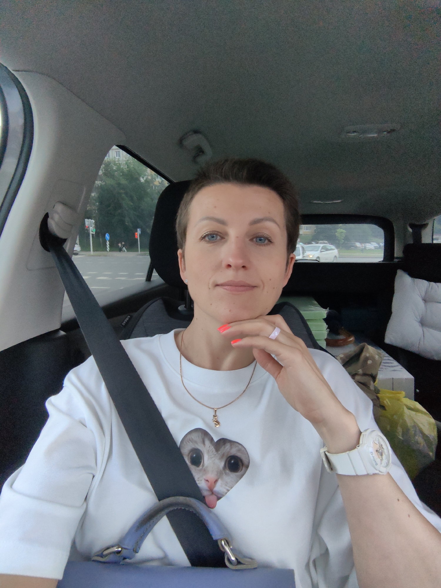 A woman with short brown hair and blue eyes is taking a selfie in the passenger seat of a car. She is wearing a white sweatshirt with a cartoon cat image on it, a gold necklace with a cat pendant, and a white watch. Her right hand is resting on her chin, and her nails are painted neon orange. A seatbelt is visible across her chest. The interior of the car is visible, including the car's ceiling, a headrest, and some items in the back seat. Through the car window, a street scene in Minsk, Belarus, can be seen, with trees, buildings, traffic lights, and other vehicles. The sky appears overcast.