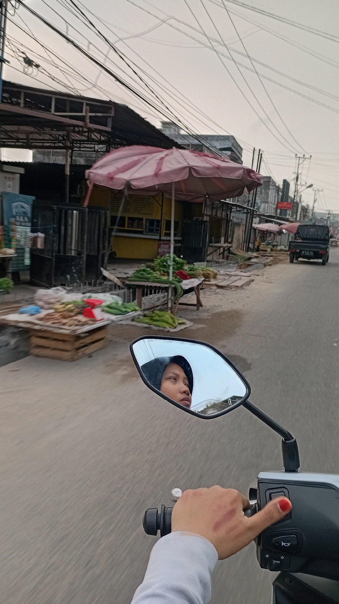 The image depicts a street scene in Jambi, Indonesia, likely a market area. The perspective is from the rider of a motorcycle, with a side mirror reflecting the face of a young person wearing a dark hijab. The person's face is turned slightly to the right, with their eyes looking forward. The background visible in the mirror shows a building with a domed roof.
In the foreground of the main image, the rider's hand is visible on the handlebar of the motorcycle, with red nail polish on the thumb. There are some red marks on the back of the hand, possibly abrasions. The motorcycle's controls are visible, including a horn button.
The street itself is paved with asphalt. To the left, there are market stalls with awnings and various goods displayed, including vegetables like corn and leafy greens on tables and in crates. Some of the stalls appear to be made of wood and metal. A yellow building with signs is partially visible behind one of the stalls. Overhead, a complex network of electrical wires stretches across the sky. In the distance, a dark-colored pickup truck is driving away from the camera, and more market stalls and buildings line the street. The sky is overcast, suggesting a possibly humid or early morning/late afternoon light. The overall impression is of a busy, everyday street scene in a Southeast Asian city.