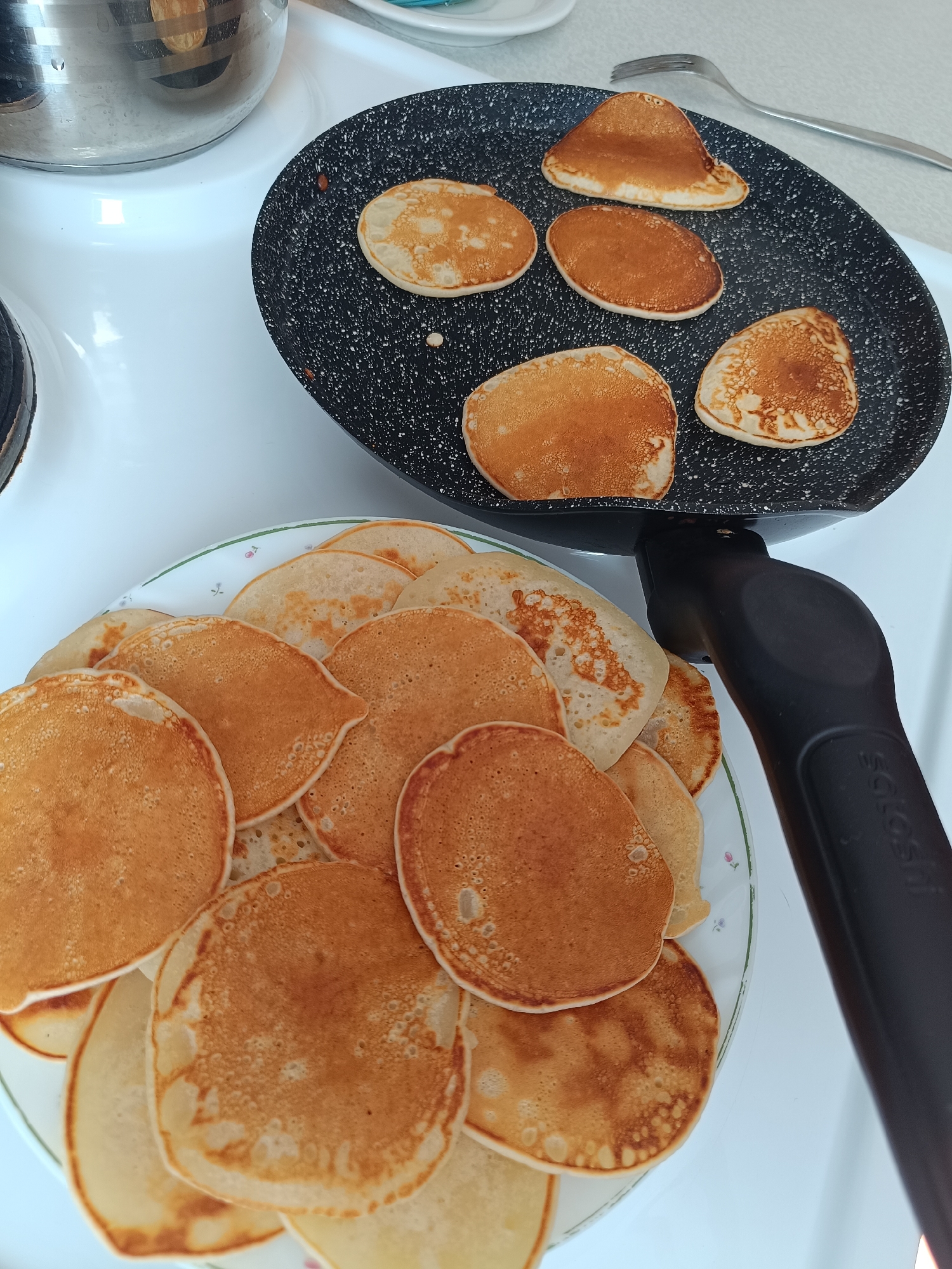 The image shows a plate of pancakes stacked on top of each other and a frying pan with pancakes cooking on a white stovetop. A stainless steel pot is next to the frying pan. A fork is near the frying pan. The setting appears to be a domestic kitchen. There is no information to suggest a connection to Tobolsk, Russia.