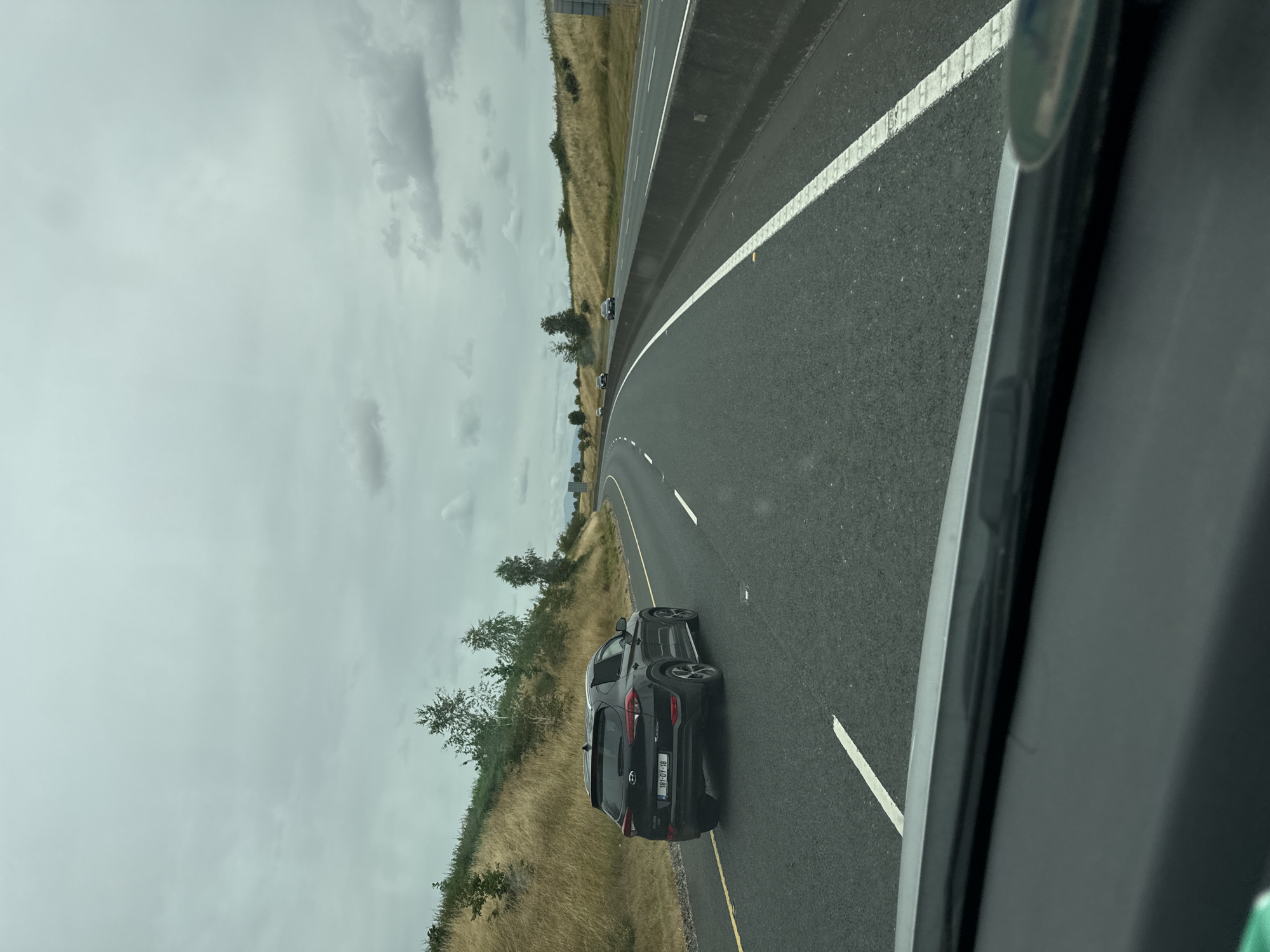 The image captures a view from a vehicle traveling on a multi-lane highway in the Moyle area of Ireland. The sky is overcast with gray clouds, suggesting a typical Irish climate. On the left side of the highway, a grassy embankment rises, dotted with sparse trees and dry, golden grass. A black Hyundai Tucson SUV, with license plate "181-OY-18", is in the left lane. The road surface is asphalt, marked with white lane dividers. On the right side of the highway, a concrete barrier separates the lanes from the shoulder, beyond which more dry vegetation and trees are visible. The highway curves gently to the right in the distance, where other vehicles can be seen. The foreground of the image shows the dashboard and windshield of the vehicle from which the photo was taken, with a hint of the car's interior.