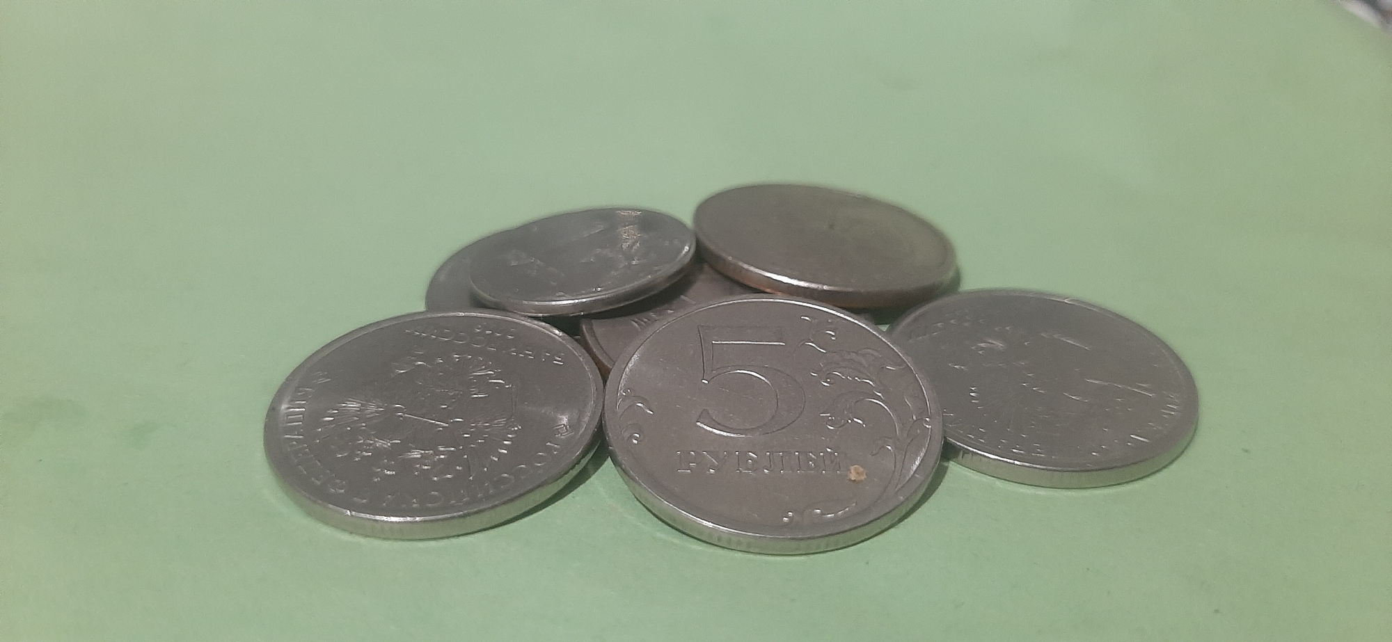 A cluster of six Russian rubles coins are arranged on a light green surface. The coins are a mixture of silver and gold tones. One coin in the foreground prominently displays the numeral "5" and the word "РУБЛЕЙ" (Rubles). Another coin to its left shows a detailed inscription and what appears to be an eagle emblem. The remaining coins are partially obscured, stacked and overlapping. The lighting suggests an indoor setting, possibly on a table or shelf. The image does not provide any information related to the city of Yessentukskaya, Russia.