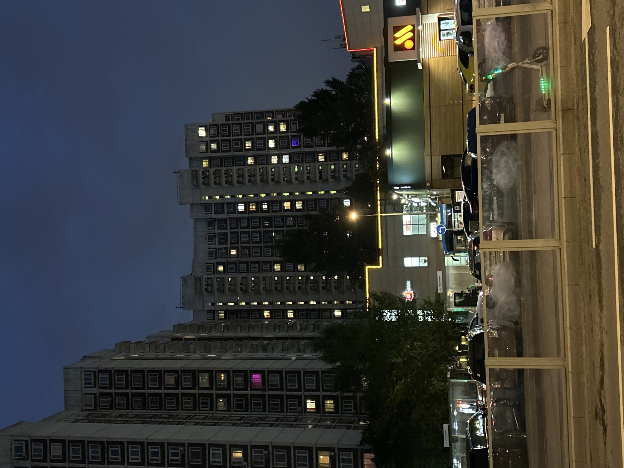 In Moscow, Russia, at night, a row of tall apartment buildings with numerous illuminated windows stands against a dark blue sky. In front of the buildings, there is a street with cars. One car is emitting a cloud of white smoke. A street light illuminates a section of the street. Further ahead, a building with a golden yellow border and the orange McDonald's logo is visible. A transparent barrier runs across the foreground, behind which a green and white electric scooter with illuminated green lights is parked. Trees are visible between the buildings and the street.