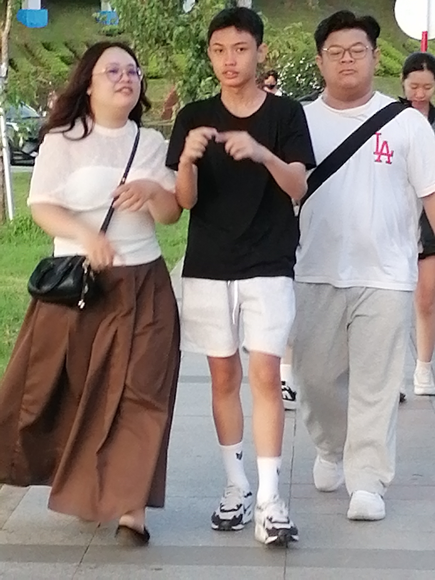 In Da Lat, Vietnam, three individuals walk on a paved walkway with a grassy area in the background. The person on the left, a female, wears a white off-the-shoulder top and a long brown skirt, carrying a black purse. In the center, a young male wears a black t-shirt and white shorts. On the right, a male wears a white t-shirt with a black and red logo and light grey sweatpants. Another female is partially visible behind the male on the right. The weather appears to be overcast.
