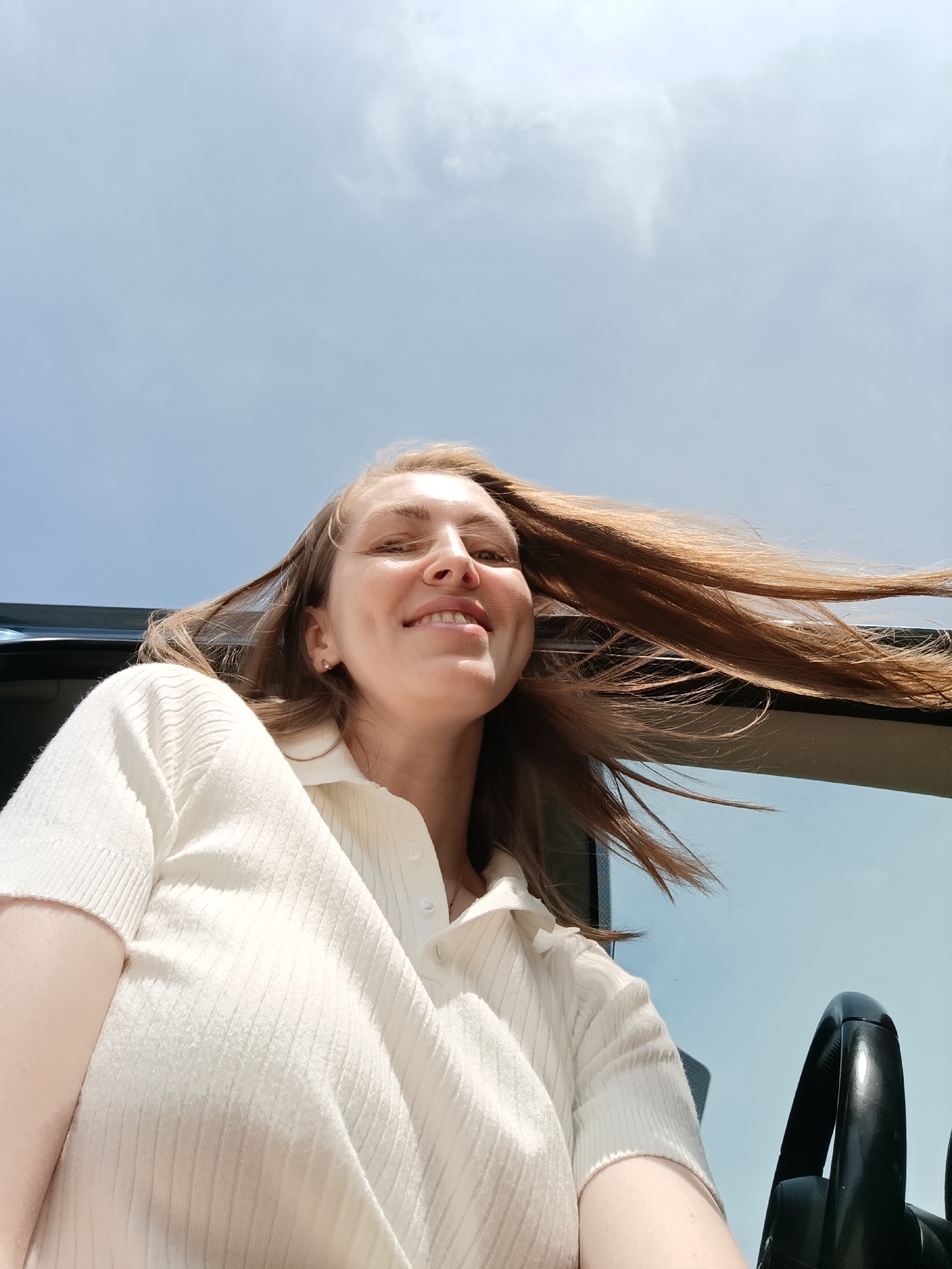 A smiling woman with long reddish-brown hair is positioned in the driver's seat of a car, likely a convertible, under a clear blue sky with scattered clouds. Her hair is blowing in the wind. She is wearing a light-colored, short-sleeved, collared shirt. A portion of the steering wheel is visible in the bottom right corner of the image. The location is not discernible to be Chaikovsky, Russia from the given image.