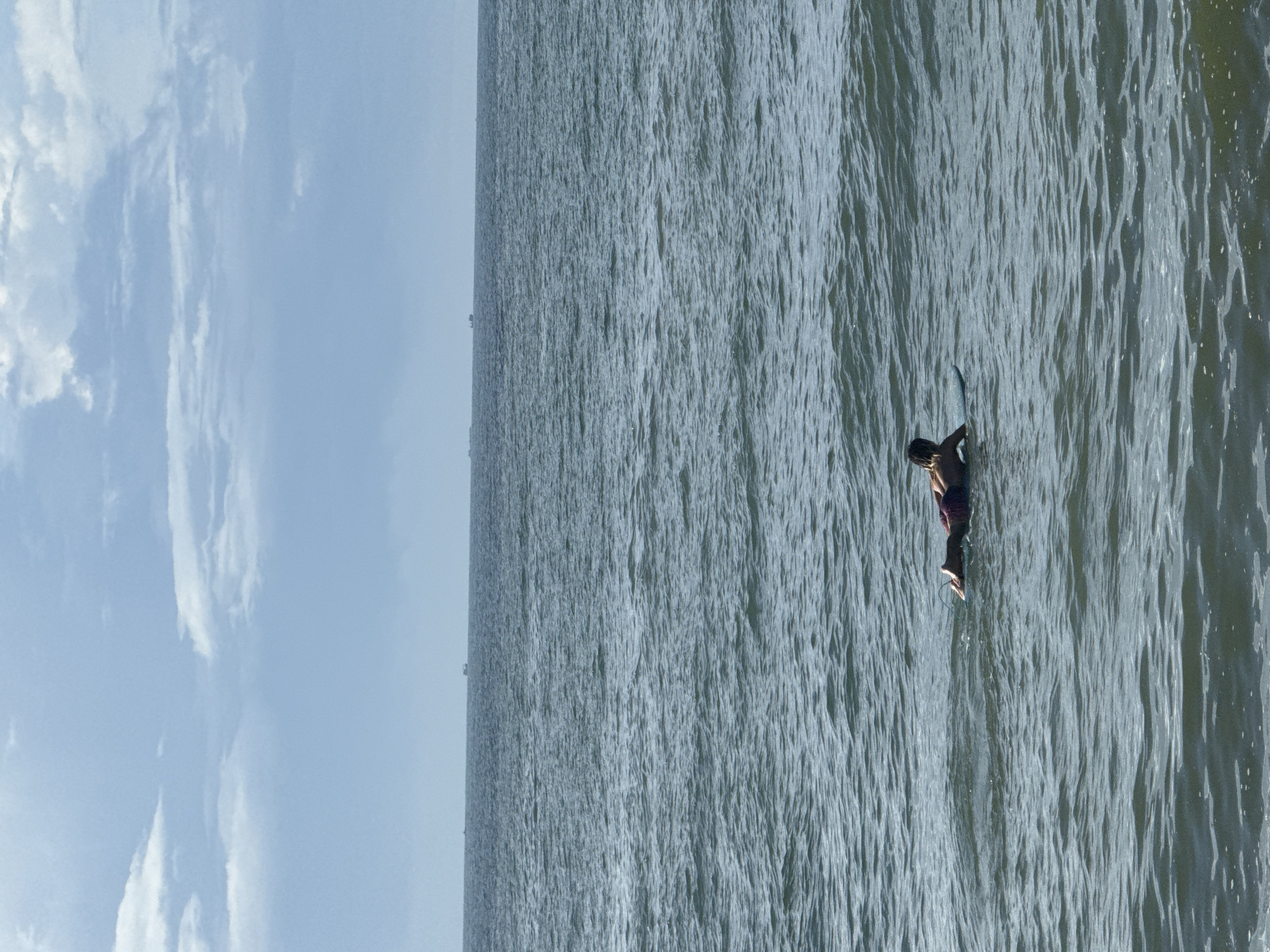 A person is lying prone on a surfboard in the ocean. The person is wearing dark swim bottoms and is positioned facing away from the camera. The surfboard is light blue. The ocean is choppy with moderate waves. In the distance, there are several small ships on the horizon. The sky is overcast with light and dark gray clouds. The image appears to be taken from a slightly elevated viewpoint, possibly a boardwalk or cliff overlooking the water. The scene is likely located at a beach in Phan Thiet, Vietnam, known for its surfing and coastal scenery.