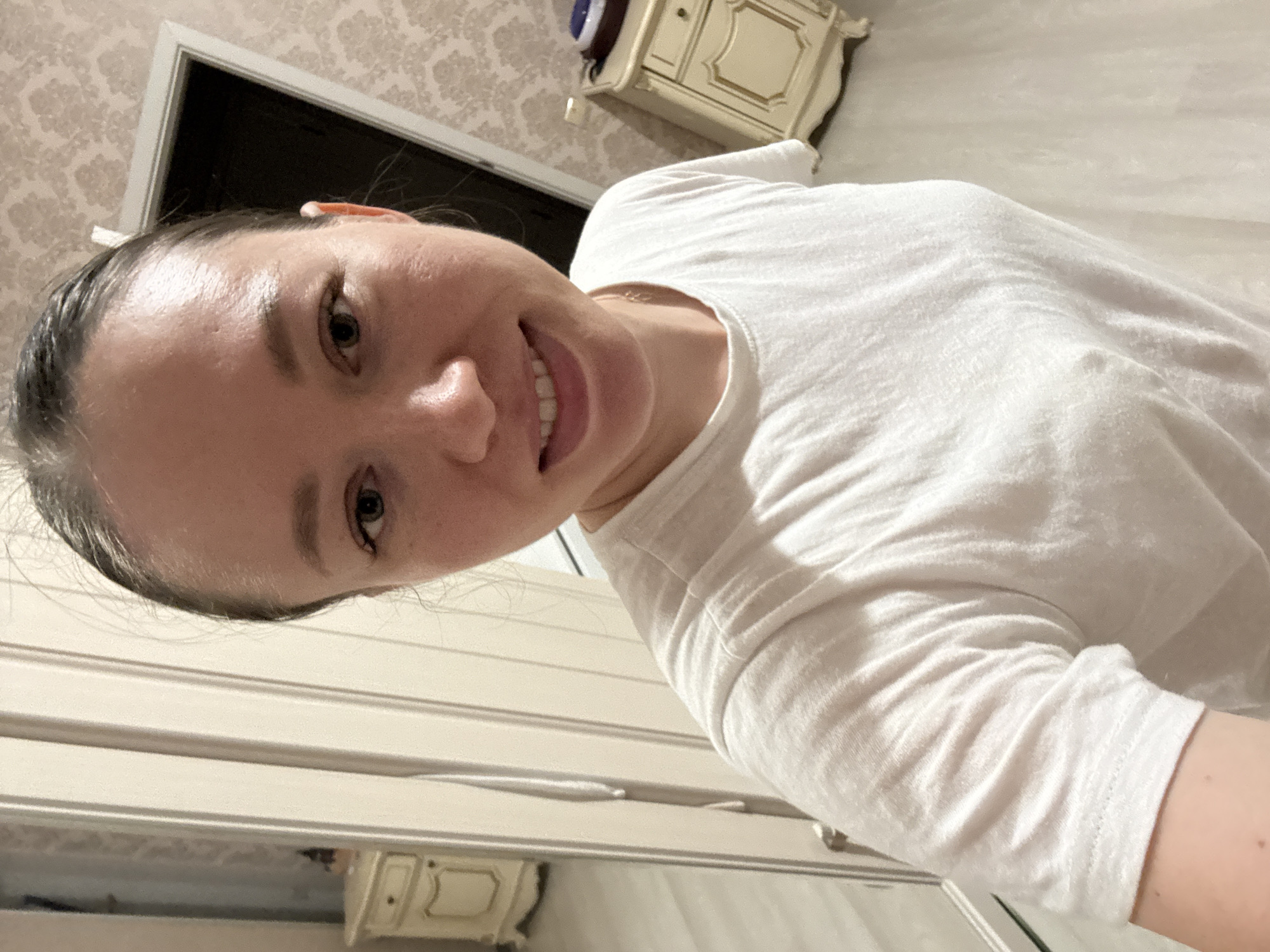 A woman with fair skin and wet, dark hair pulled back smiles at the camera. She is wearing a white long-sleeved t-shirt. The background includes a bedroom or living room with light-colored walls that feature a patterned wallpaper design. To the left are light-colored cabinets. In the upper right corner, a white and gold nightstand is visible, with a small blue object on top. The scene appears to be indoors. There is no evidence in the photo to indicate the exact location of Ферма, Russia.