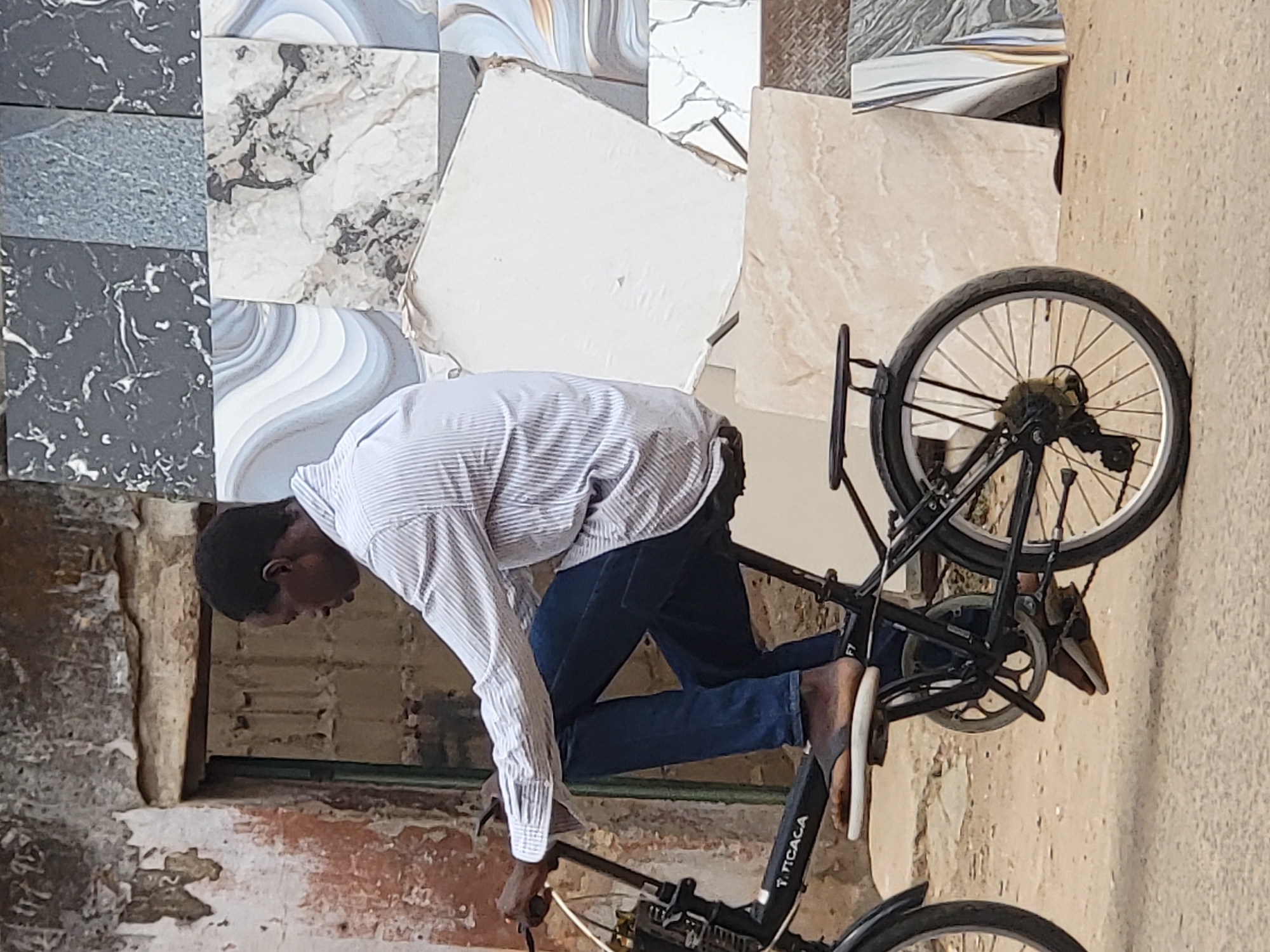 A man in a striped shirt and jeans bends over a bicycle in Maiburiji, Nigeria. The bicycle is black and appears to be resting against a wall decorated with tile samples. The wall behind the man and bicycle appears to be constructed of stone and brick.