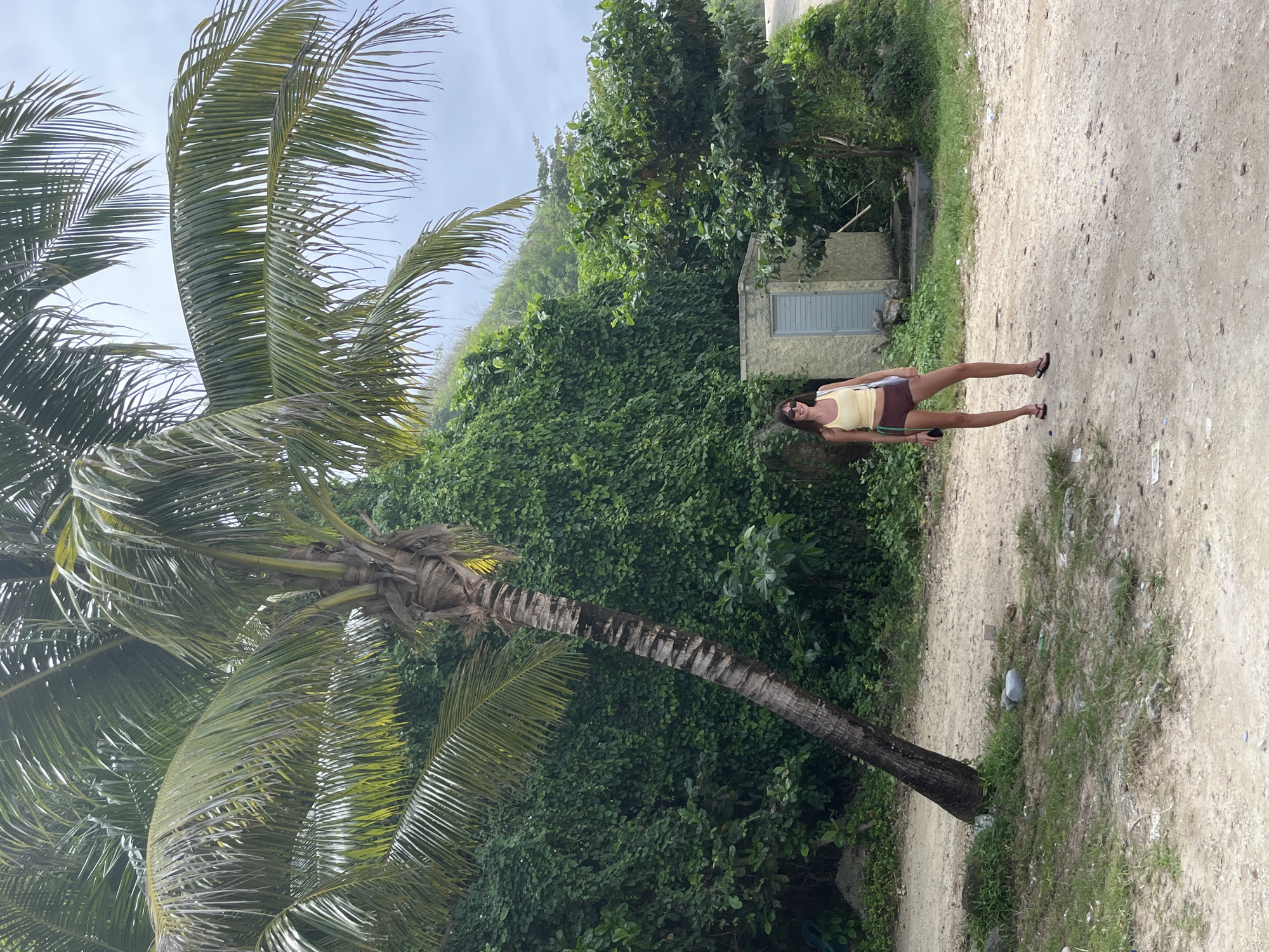 A woman stands on a dirt road near a palm tree in Kuta Selatan, Indonesia. She wears a yellow tank top, brown shorts, and black sandals. The background features lush green vegetation and a small, nondescript building. The sky is overcast.