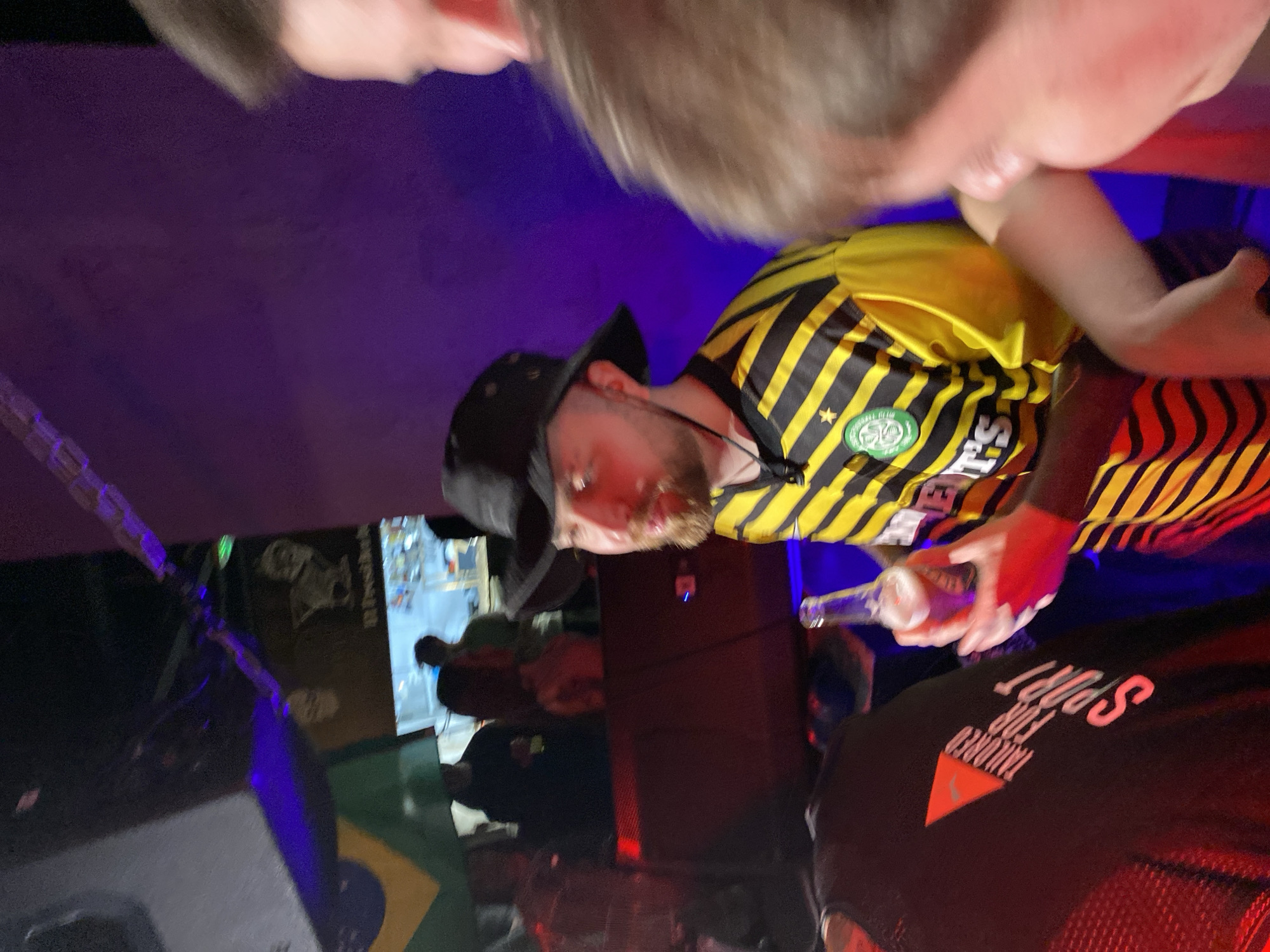 The image depicts a group of people in what appears to be an indoor setting with a bar visible in the background. One man in the center is wearing a black bucket hat, a yellow and black striped soccer jersey, and holding a glass bottle. Another person, perhaps posing for a photo, is partially visible at the top of the frame. The location might be a bar in Izhevsk, Russia, considering the text on the black shirt of another person says "TAILORED FOR SPORT," and the colors of the Brazil flag (green and yellow) are present on the floor. The lighting is somewhat dim and colorful, with blue and red hues dominating the scene. Several more people are visible towards the background near what could be the bar area, but their actions and interactions are not clear. The focus of the image is primarily on the man wearing the soccer jersey.