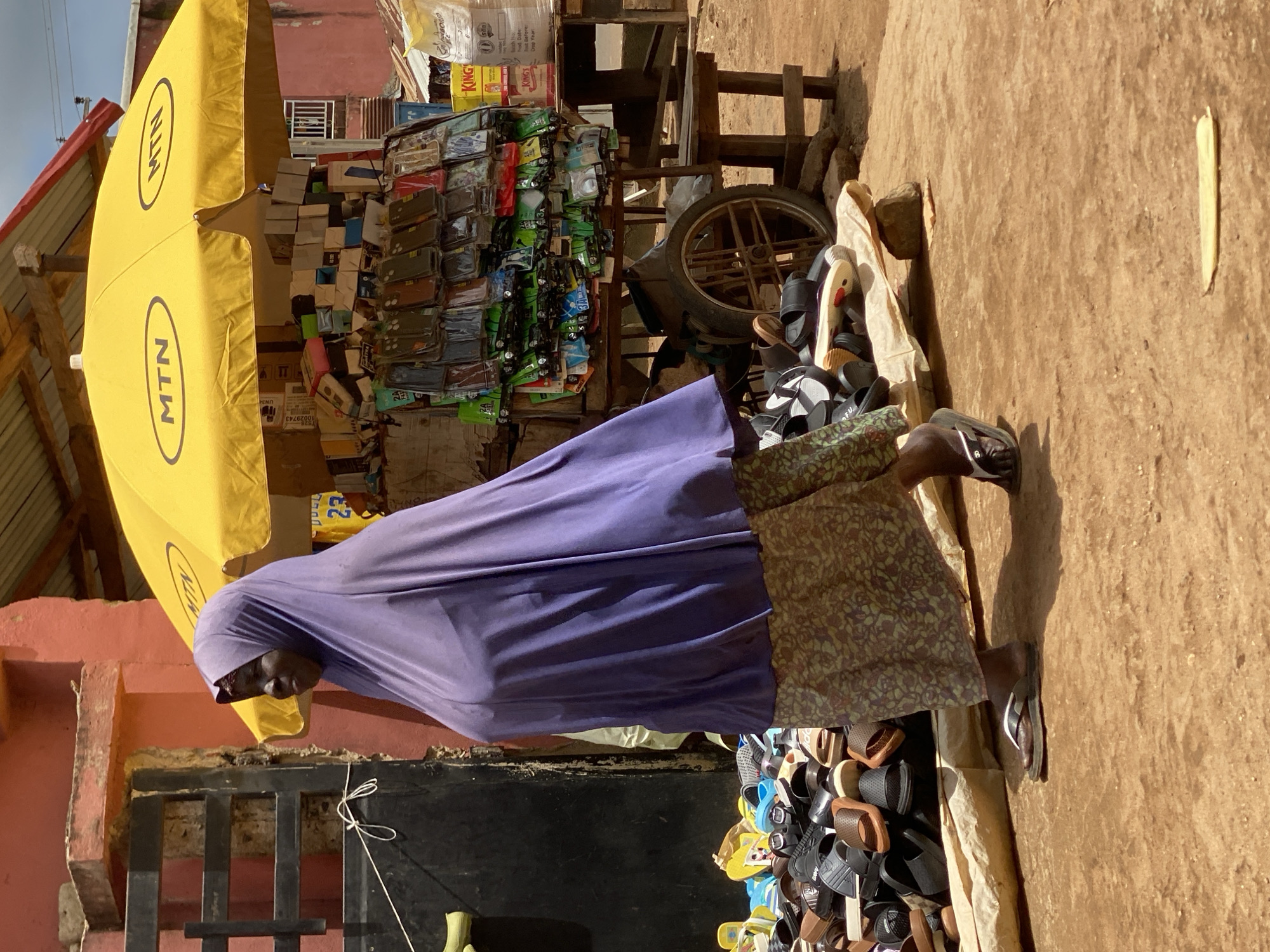 The image depicts an outdoor market scene in the city of Kafanchan B, Nigeria. A woman wearing a purple hijab and a patterned skirt walks across a dirt ground. She is wearing flip-flops. To her right, a yellow umbrella with "MTN" logos is set up, shading a stall displaying numerous cell phone cases and accessories. Behind the stall, a cart with a large wheel is visible. A pile of sandals and flip-flops is laid out on the ground in front of the stall. The background shows a reddish building and more market stalls with various goods. The scene suggests a busy commercial area.