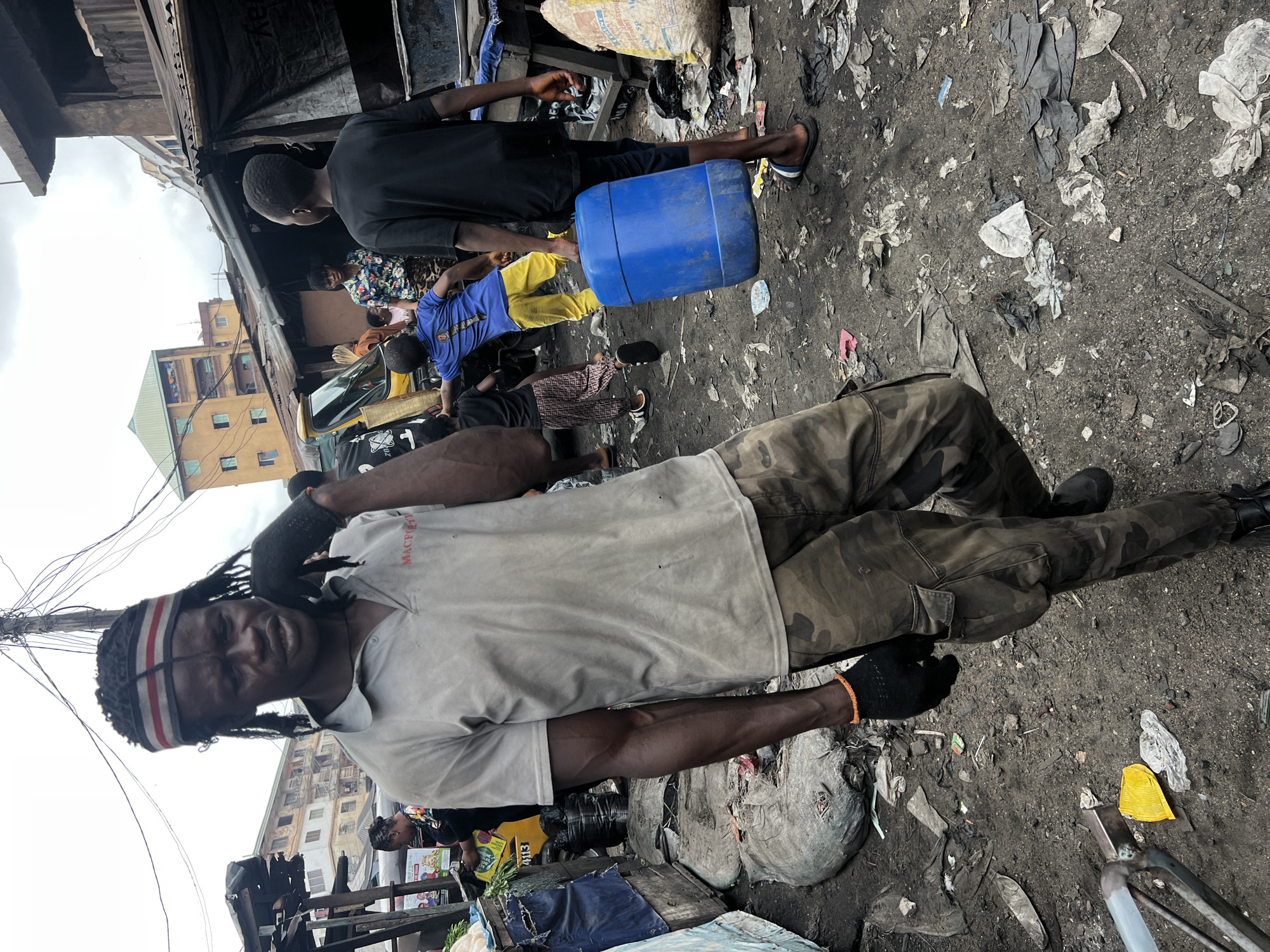 The image captures a bustling street or market scene in Nkpor, Nigeria, under an overcast daytime sky. The ground is uneven, made of dirt and debris, including plastic bags, paper, and fabric scraps.
In the foreground, a dark-skinned man with dreadlocks and a red, white, and black headband looks directly at the camera. He wears a light-colored short-sleeve t-shirt with "MACPREM" printed on the left chest, camouflage cargo pants, black boots, and black gloves on both hands. His right arm is bent, holding a dark, possibly heavy, object or material against his shoulder.
Several other individuals populate the mid-ground and background. To the right of the main subject, a person in a black t-shirt and shorts stands near a blue plastic barrel lying on its side. Beyond them, another person is seen in a blue shirt and yellow pants, possibly crouching. Further back, a woman in a floral-patterned top is visible among other less distinct figures.
The background features improvised market stalls or shelters constructed from corrugated metal and tarps. Behind these structures, multi-story buildings, characteristic of urban or semi-urban dwellings, are visible, some with laundry hanging from balconies. A tangle of black electrical or communication wires crisscrosses overhead in the upper left. A white sack, possibly filled with goods, stands upright near the blue barrel, and a yellow box with "4113" printed on it is discernible in the mid-ground. The scene conveys a sense of daily life and commerce in a lively, informal setting.