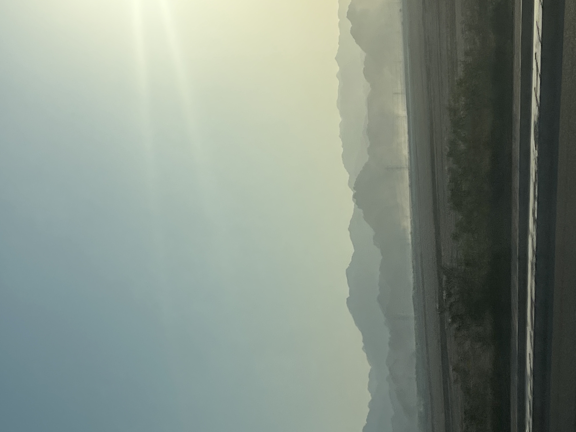 The image depicts a vast, hazy mountain range in the distance, silhouetted against a pale blue and yellow sky. The mountains are layered, with the furthest peaks appearing faint and indistinct, suggesting a considerable distance. In the foreground, there is a flat, dark expanse, possibly agricultural land or a barren desert area, with sparse vegetation. A paved road or pathway runs along the bottom of the frame, with a reflective surface. There are no visible people or animals. The overall impression is one of a wide, open, and somewhat desolate landscape, possibly taken during a time of day with low light or atmospheric conditions like dust or mist. The scene is likely located in the desert region surrounding Sharm El Sheikh, Egypt, known for its mountainous terrain and arid environment.