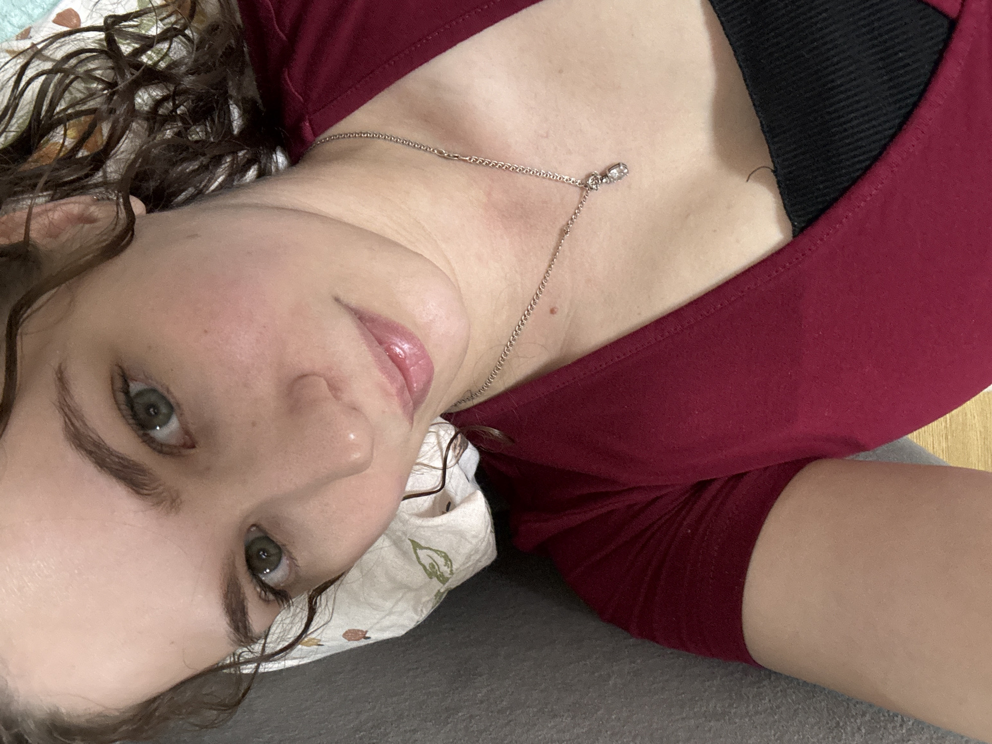 A close-up, high-angle selfie captures a person, likely female, lying down indoors, possibly on a couch or bed in Chaikovsky, Russia. The subject's head is tilted, with their gaze directed upwards towards the camera. They have fair skin, green eyes, and dark, curly hair that rests on a light-colored pillow with a subtle leaf or floral pattern. A faint, natural smile is visible on their lips, which appear glossy and pink.
The person is wearing a maroon-colored top or dress, beneath which a black ribbed undergarment is partially visible at the neckline. A delicate silver chain necklace with a small, clear gemstone pendant hangs around their neck. The background consists of the grey upholstered surface they are resting on and a glimpse of what appears to be a light brown or wooden surface on the right side. The overall impression is one of calm or relaxation. No text is visible in the image.