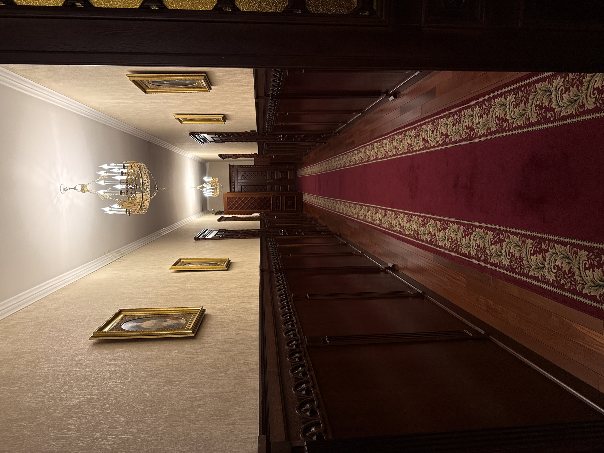 The image shows a long hallway in what appears to be an ornate interior. A red carpet with a floral border runs the length of the hallway. Dark wood paneling lines the lower portion of the walls on either side. Above the paneling, the walls are a textured beige color with gold-framed paintings spaced along the wall. Chandeliers hang from the ceiling, spaced out along the hallway. Based on the architectural style, ornate details, and a general historical appearance, it is likely a hallway in a building in the city of Saint Petersburg, Russia. No people are visible in the image.