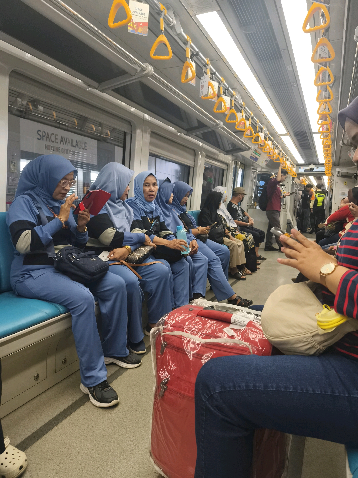 The image captures the interior of a train carriage, likely part of the transportation system in Palembang, Indonesia, as suggested by the context. Several individuals are seated, predominantly women wearing blue uniforms and headscarves, suggesting a group traveling together, possibly for work. One woman in the foreground is engrossed in her tablet, while others appear to be conversing or resting. The carriage is equipped with standard amenities, including yellow safety handles for standing passengers and overhead lighting. Luggage, including a prominent red suitcase covered in plastic, is visible, indicating a journey. The overall atmosphere is that of ordinary public transport, with passengers engaged in their individual activities during their commute.