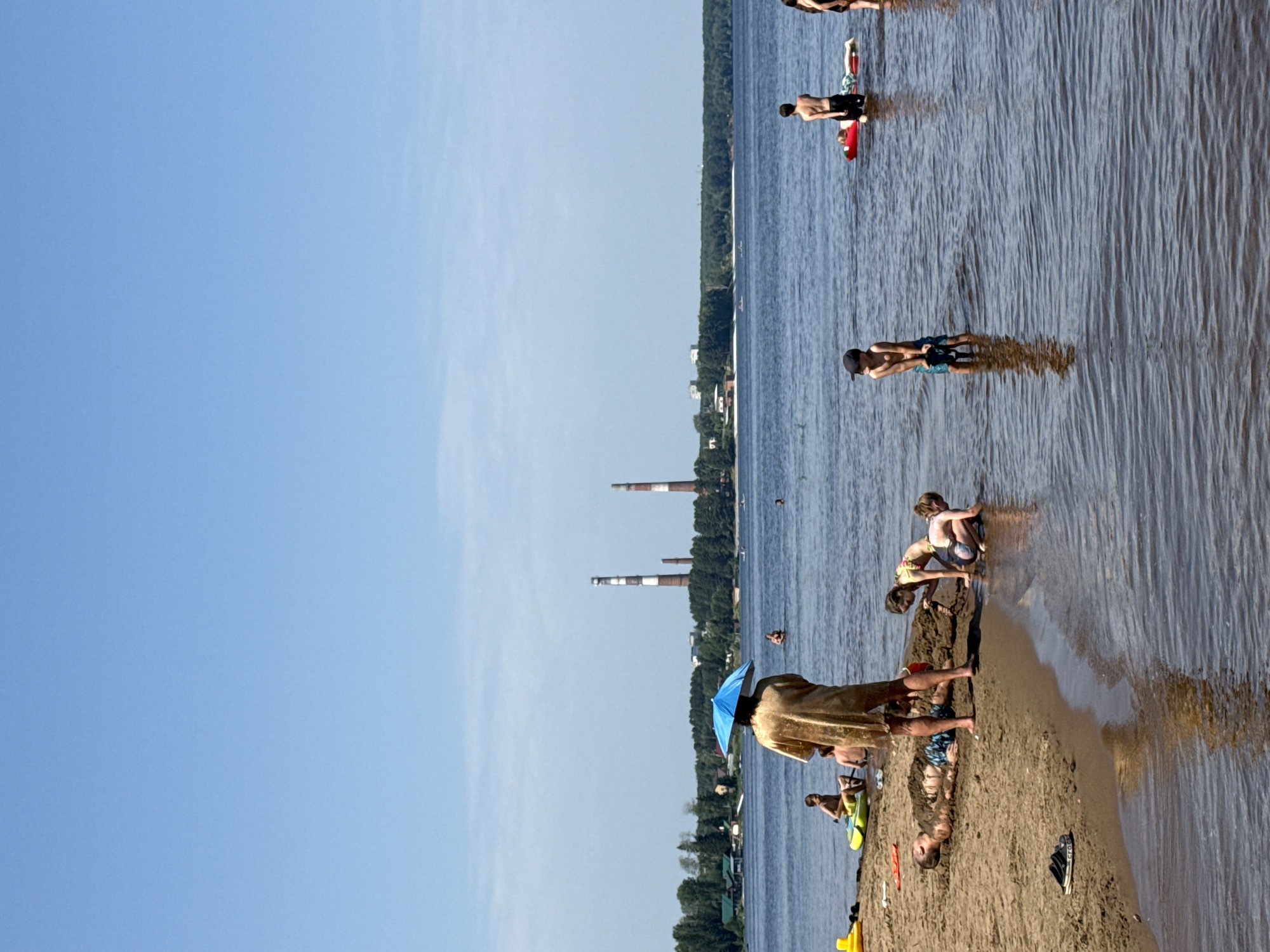 A group of people are enjoying a sunny day at a beach with a body of water in the foreground. The water is calm and reflects the blue sky. In the background, there is a treeline with some buildings and industrial smokestacks visible.
On the sandy shore, a person stands facing away from the camera, holding a blue umbrella over their head, and wearing a tan robe. To the left of this person, a man is lying on the sand, partially covered in it, with a pair of black flip-flops beside him. Two children, a girl and a boy, are playing in the sand near the man.
In the water, several children are visible. One boy is standing in the water, wearing a black cap and blue shorts. Another boy is sitting on a yellow inflatable raft. Further in the water, a person is standing on a red inflatable raft. Another person is standing in the water in the distance.