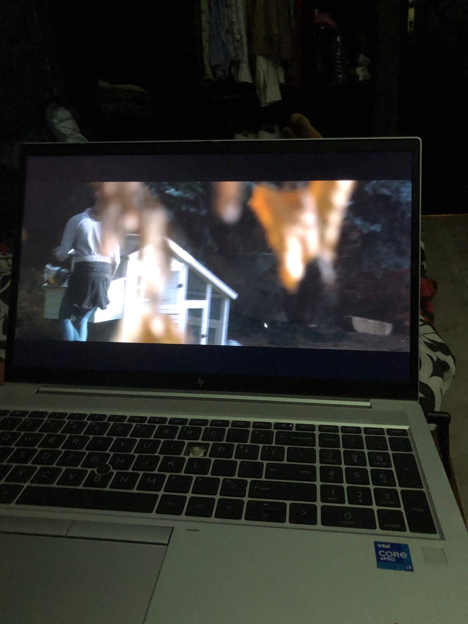 A laptop is placed on a patterned surface. The laptop screen displays a scene with a person wearing a light-colored shirt and a dark skirt, standing next to a small, white structure resembling a playhouse. The background of the screen shows dark foliage. The laptop keyboard is visible below the screen, with a portion of the trackpad to the left. An "Intel Core i7" sticker is present on the lower right of the laptop. The surrounding environment is dimly lit, suggesting an indoor setting at night. There are no visible landmarks or clues that would indicate the location in Freetown, Sierra Leone.