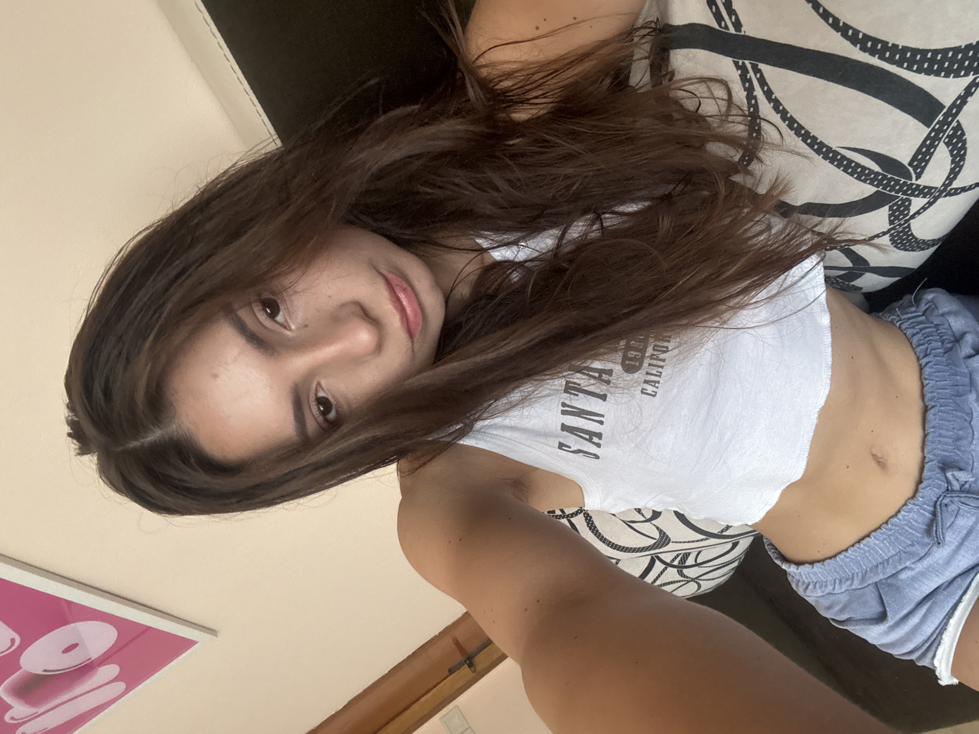 This is a selfie taken by a young woman with long, dark brown hair. She is lying down, possibly on a sofa or bed, as indicated by the pillow with a black and white chain-link pattern behind her and the dark surface beneath. She is wearing a white crop top with the text "SANTA MONICA CALIFORNIA" printed on it in black lettering, and light blue drawstring shorts. Her expression is relaxed and she appears to be looking directly at the camera with a slight smile.
The setting appears to be indoors, with a wall painted a light, creamy color visible in the background. A pink poster with abstract circular shapes is partially visible on the wall in the lower left corner of the image. The lighting suggests it is daytime, but no specific time of day can be determined. The overall mood is casual and intimate. The image was taken in Varna, Bulgaria.