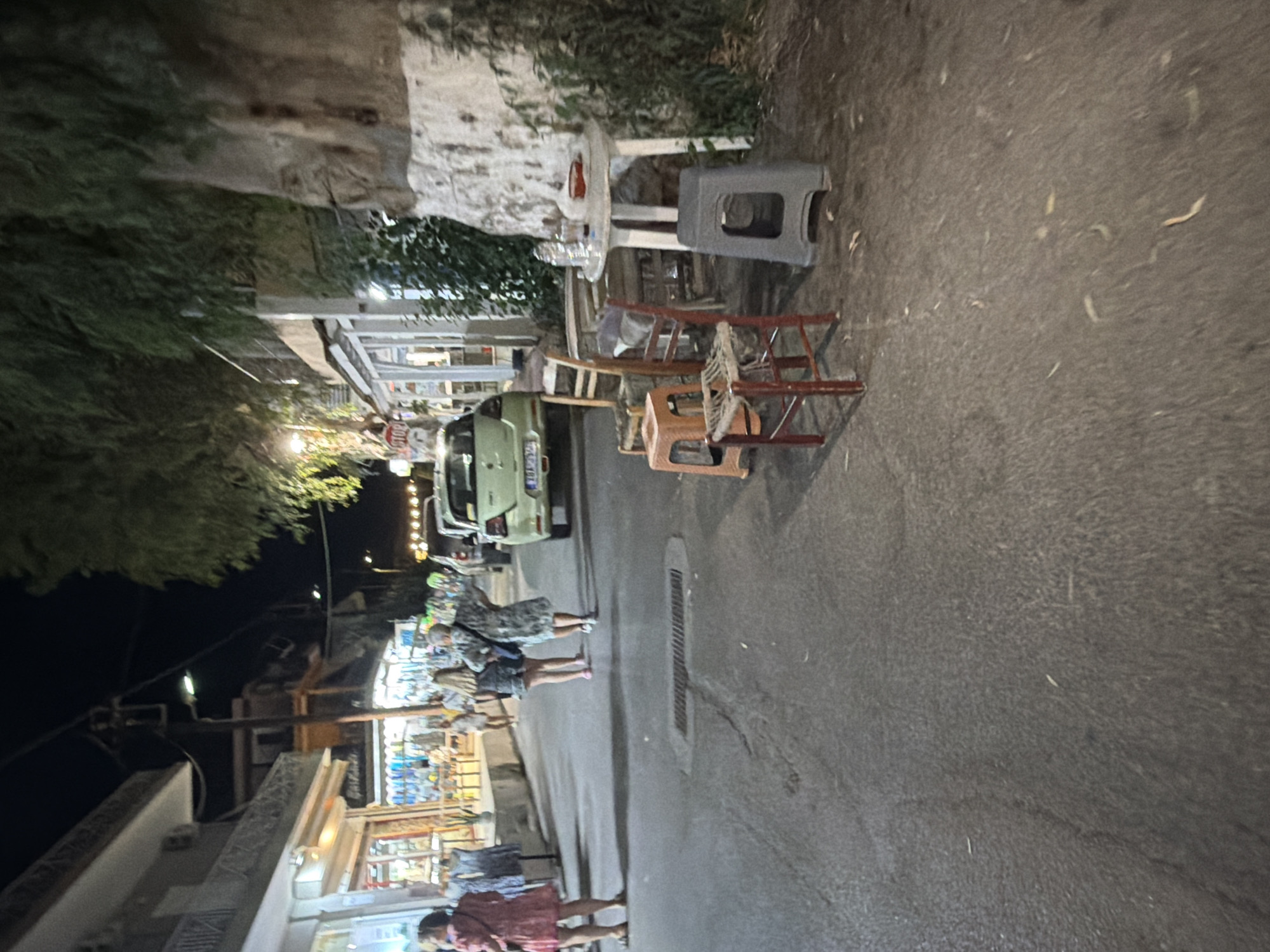The image depicts a nighttime street scene in Georgioupoli, Greece. On the left side of the street are shops with illuminated interiors displaying merchandise. Several people are walking along the sidewalk and street. One woman in a pink dress is standing near a shop entrance. Further down the street, two women in casual attire are walking away from the camera. On the right side of the street, a light green car is parked. In the foreground, there are several chairs and a small white table set up outside what appears to be a cafe or restaurant, with a few items on the table. The street is paved, and there is some greenery and a rocky cliff face in the background. The overall impression is of a casual evening in a Greek coastal town.