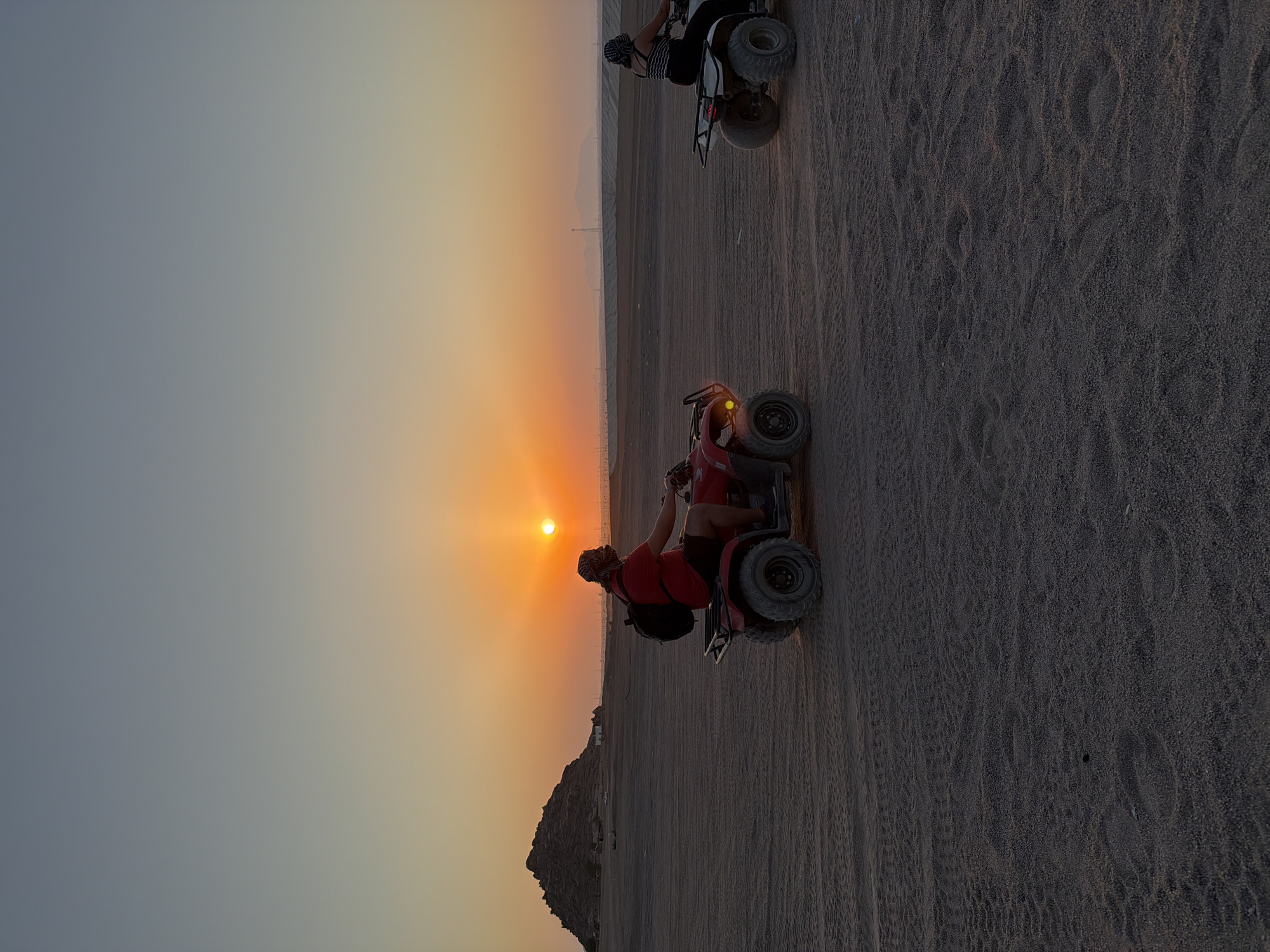 Two individuals are riding quad bikes in a desert landscape at sunset. The person on the left is riding a red quad bike and is wearing a red shirt, dark shorts, and a headscarf. They have a black backpack. The person on the right is riding a white quad bike and is wearing a striped top and dark pants, also with a headscarf. The sun is low on the horizon, casting a warm orange glow across the sky and reflecting off the sand. In the background, there is a rocky hill formation on the left and a flat, arid plain stretching to the horizon with some distant structures and power lines. The ground is sandy with tire tracks visible. The scene depicts an outdoor recreational activity in a desert environment, likely in Sharm El Sheikh, Egypt, known for its desert landscapes and sunset activities.