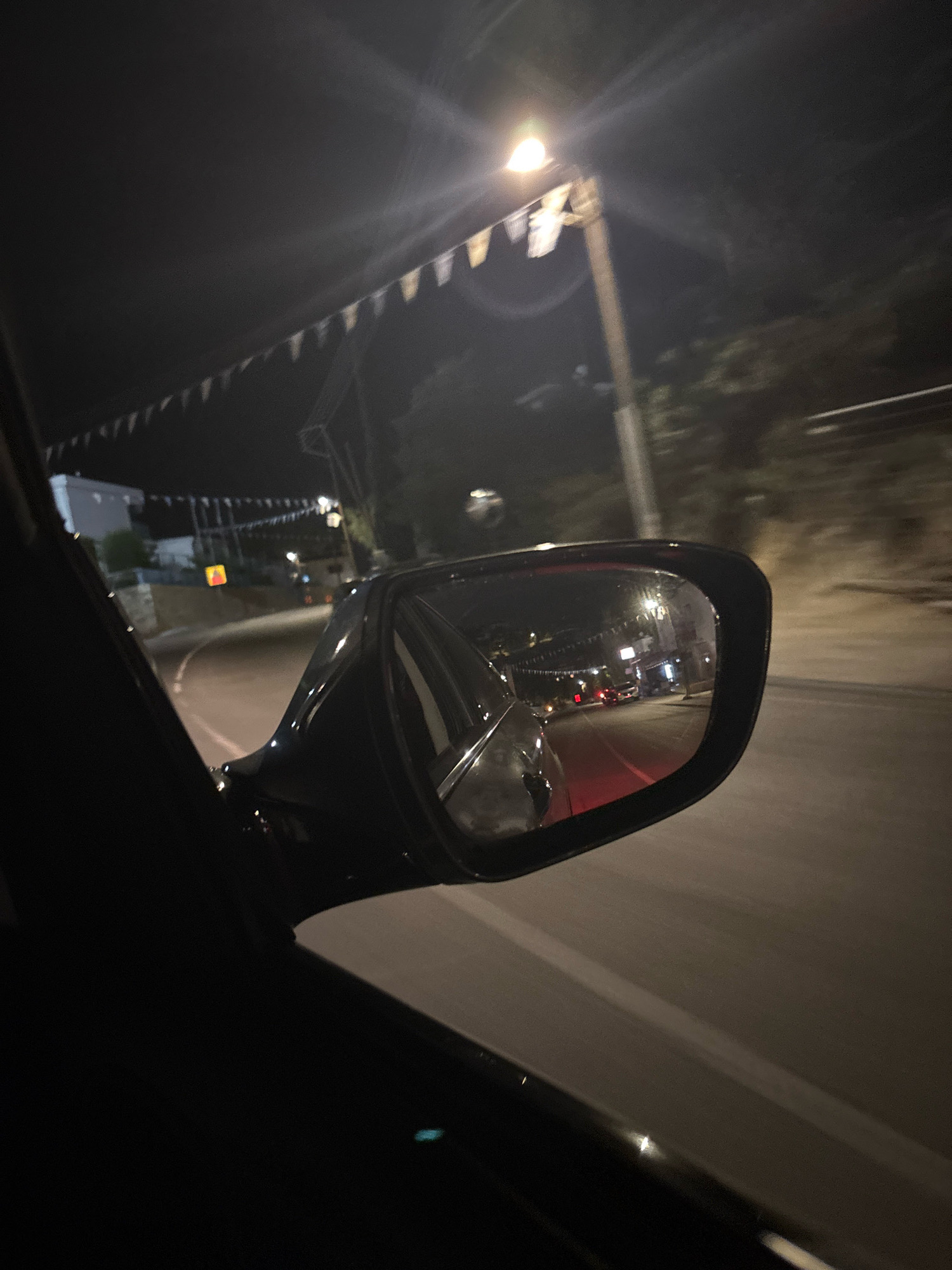 The image captures a nighttime view from inside a car, looking out towards the side and into the wing mirror. The car is on a road in Agios Tychona Community, Cyprus, as indicated by the presence of streetlights, buildings, and foliage consistent with a Mediterranean coastal town.
In the foreground, the dark silhouette of the car's interior is visible, including the door frame and the side of the car. The wing mirror, positioned on the right side of the frame, reflects the scene behind the car. This reflection shows a road with faint lane markings, illuminated by streetlights and the headlights of other vehicles. Several cars are visible in the distance, moving along the road.
The background outside the car, to the left, shows buildings with lights on, suggesting a residential or commercial area. A string of triangular flags, possibly decorative for an event, stretches across the upper portion of the frame, illuminated by a bright streetlight. The overall atmosphere is dark, characteristic of a night drive. The road surface appears to be asphalt. There are no identifiable individuals clearly visible.