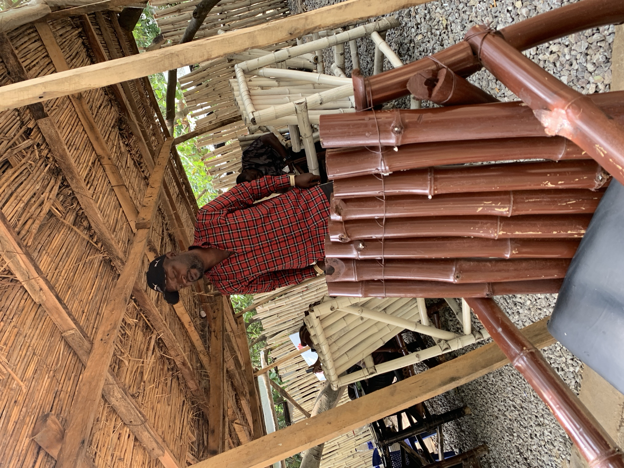 The image depicts an outdoor seating area, likely a restaurant or bar, with a rustic ambiance, constructed primarily from bamboo and natural materials. The roofing is made of thatched straw supported by wooden beams, creating a sheltered yet open-air environment. In the foreground, a dark-colored chair made of painted bamboo is visible, with its legs and backrest constructed from thick, polished bamboo poles.
A man, wearing a red and black plaid shirt and a black baseball cap, stands in the middle ground, looking downwards. He has a beard and is wearing a gold watch on his left wrist. Behind him, other bamboo furniture, including chairs and tables, are arranged. Another man is seated in the background, wearing a patterned shirt. The ground is covered with gravel. The overall setting suggests a casual, outdoor establishment in a tropical or semi-tropical climate. Based on the visual cues and common architectural styles in the region, this scene is consistent with an outdoor cafe or bar located in Abuja, Nigeria.