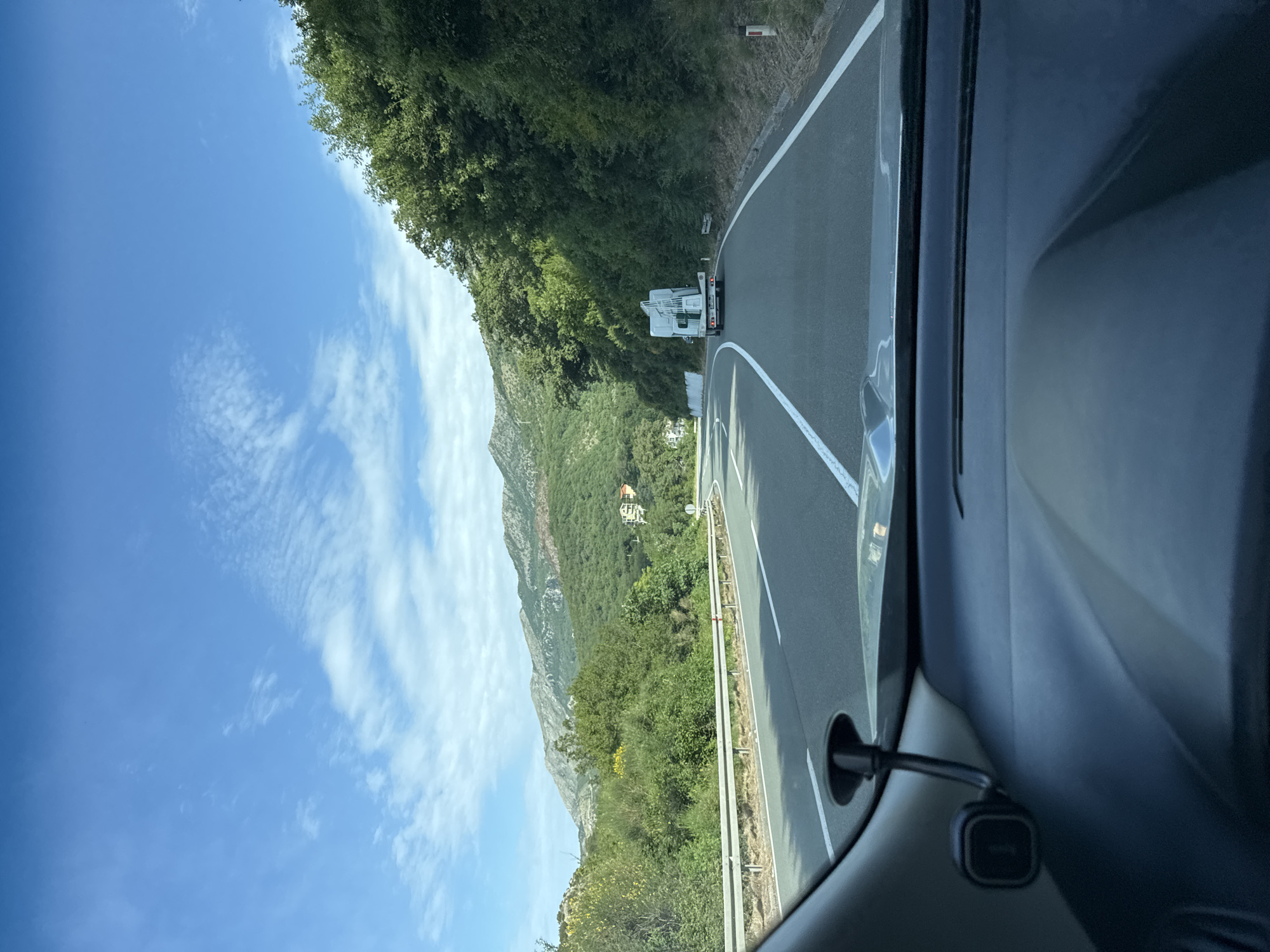 The image depicts a view from inside a vehicle driving on a winding road near Golubovići, Montenegro. The road has solid white lines delineating the lanes. In the distance, a white truck is visible on the road ahead. A mountain range covered in green vegetation is visible in the background, along with a clear blue sky with scattered clouds. Part of the vehicle's interior, including the dashboard and a small electronic device mounted on it, are in the foreground.