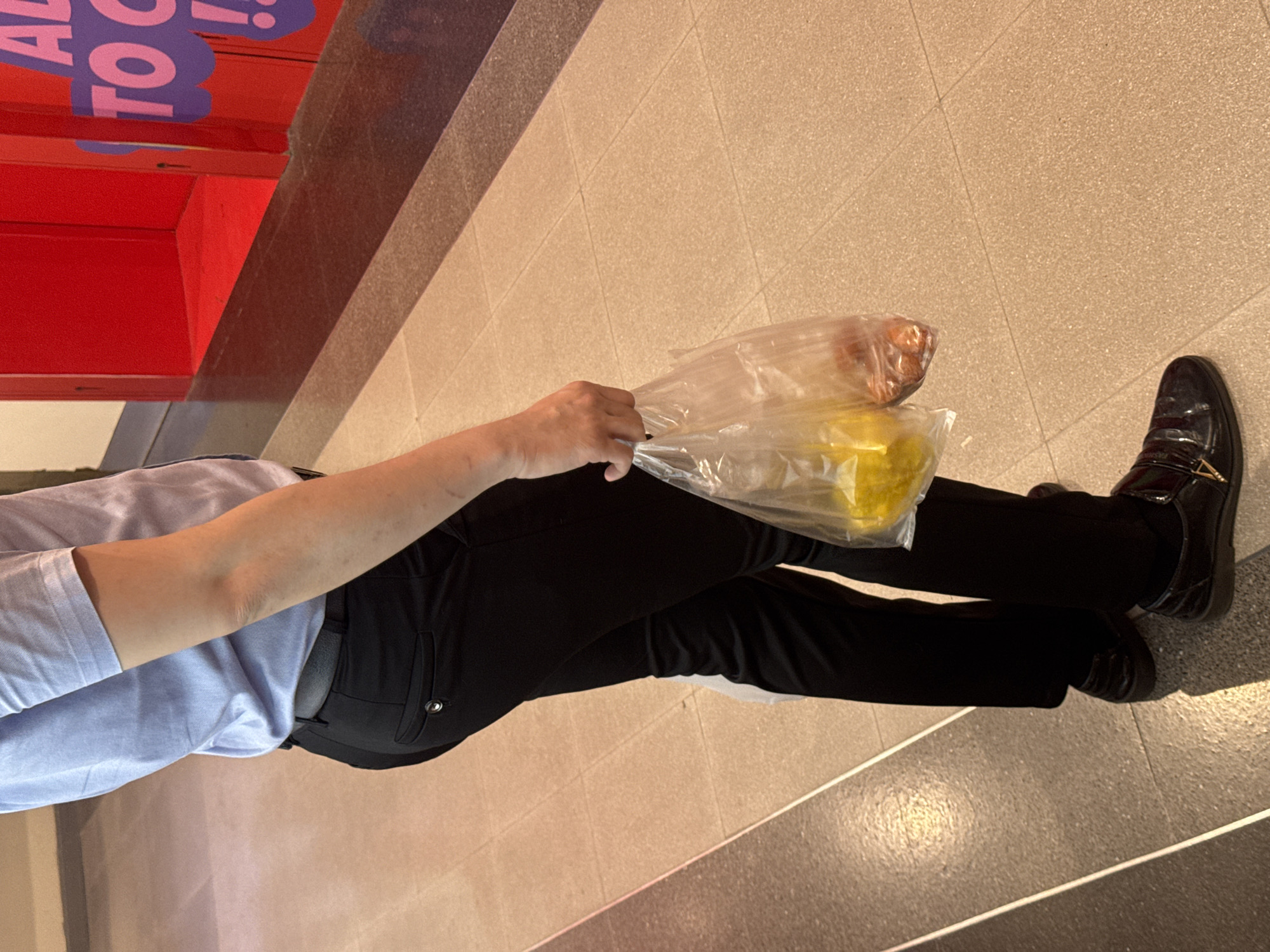 A person is standing on a light-colored tiled floor in what appears to be a public indoor area. The person is wearing a light blue short-sleeved collared shirt, black trousers, and black dress shoes. They are holding two clear plastic bags in their right hand. One bag contains yellow items, and the other contains small reddish-brown items. In the background, a bright red structure with purple lettering is visible. The scene suggests a person who has made a purchase. There is no visible information to pinpoint the location as Nong Pa Khrang, Thailand.