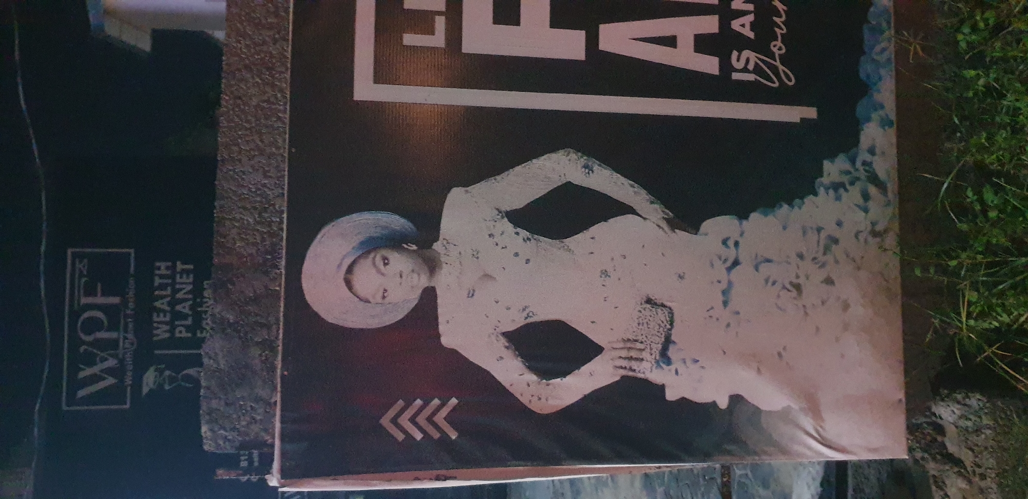 A banner advertising "Wealth Planet Fashion" is displayed outdoors at night in Narayi, Nigeria. The banner features a woman in a white, ornate dress and a large, circular hat. She is holding a clutch bag. To the right of the woman, large white text reads "LIF" and partially visible text below reads "IS AN" and "Journ". The banner is mounted on a rough, textured surface, possibly a wall or fence, with some greenery visible at the bottom. The overall lighting suggests it is nighttime.