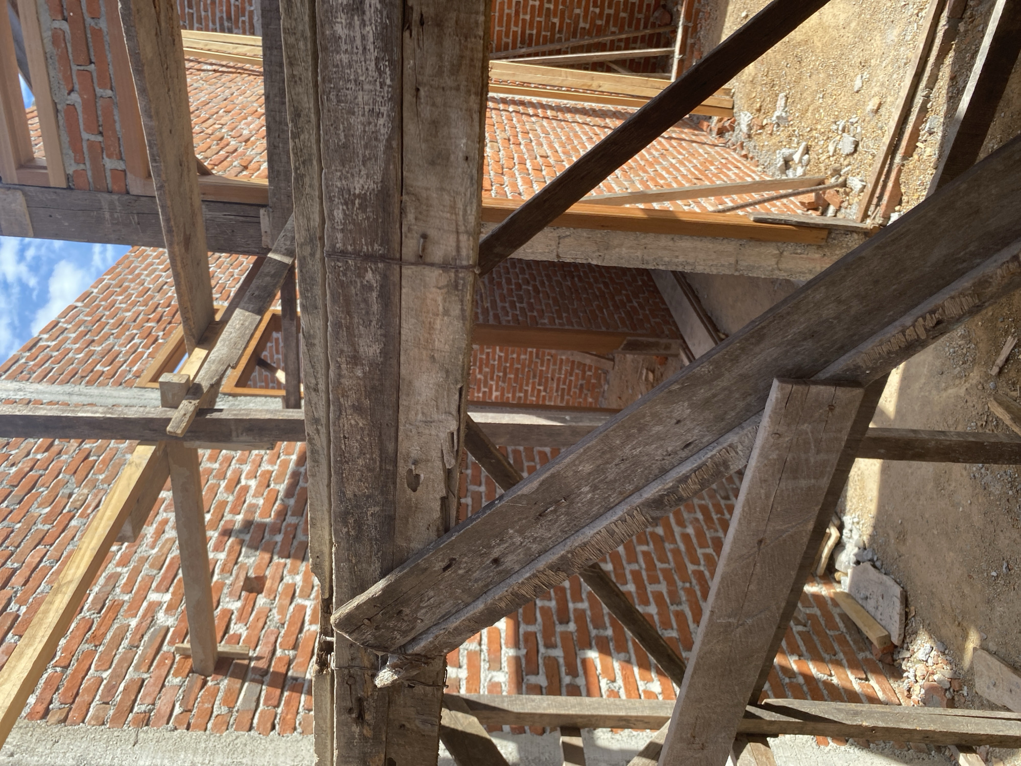 The image shows the interior of a brick building under construction, with wooden scaffolding in the foreground. The walls are made of red bricks, and there are wooden beams and supports visible throughout the scene. The ground is covered in dirt and rubble. The sky is visible in the upper right corner, with some clouds. The lighting suggests it is daytime.