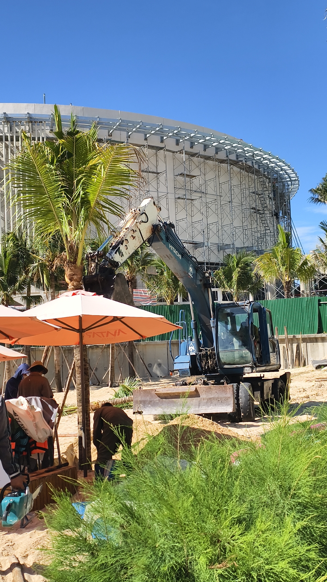 An excavator is shown on a sandy construction site in Nha Trang, Vietnam, in front of a large building under construction. The building is partially covered in scaffolding, and its curved facade is visible. Several palm trees are scattered across the scene. Two orange umbrellas are open, providing shade for a group of people who are gathered on the sand. One person is operating the excavator, while others appear to be involved in construction-related activities or taking a break. The ground is covered in sand and some patches of greenery. The sky is clear and blue.