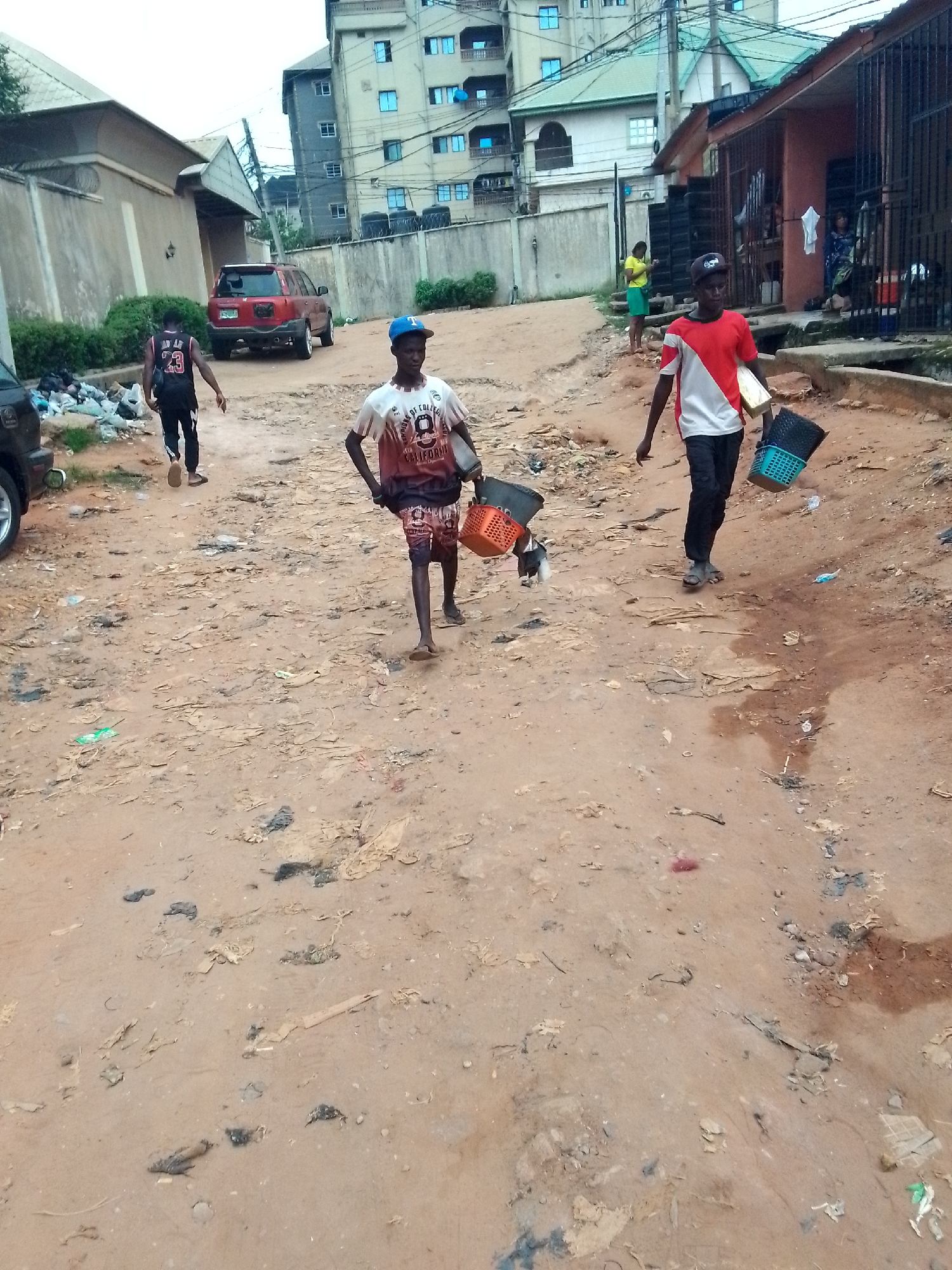 The image captures a street scene in Nkpor, Nigeria. The road is unpaved, covered in a layer of reddish-brown dirt and scattered with trash. Three young men are walking along the road. One wears a blue baseball cap, a t-shirt with a graphic print, and shorts, and carries orange and black buckets. Another man wears a red and white t-shirt, dark pants, and a baseball cap, and carries a blue and black plastic basket. The third man is wearing a number 23 basketball jersey and dark pants. Behind them, a red SUV is parked near a building. Several other buildings are also visible. Two people are visible in front of a building with a metal fence. Another person stands near the building's gate.
