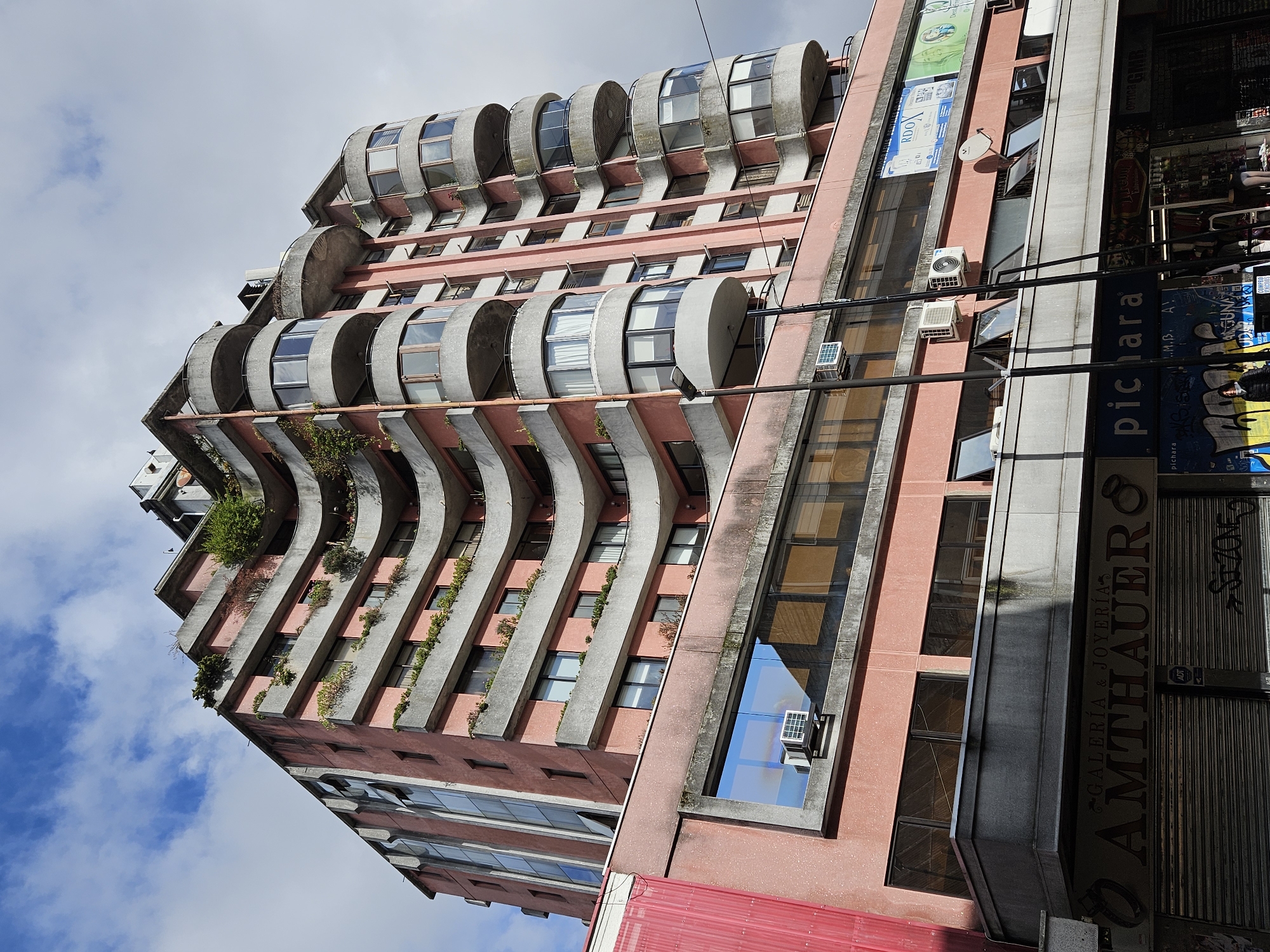 This image captures a multi-story building in Osorno, Chile, showcasing a distinctive architectural style under a partly cloudy sky. The building features a salmon-pink (or terracotta) colored facade contrasted with prominent, raw concrete elements. Its most striking feature is a series of undulating or wave-like concrete structures that project outwards, forming balconies. Each of these balcony sections encloses cylindrical or barrel-shaped windows. Green plants and vines grow along several of these concrete ledges, adding a touch of nature to the urban structure.
The building's lower section on the right side appears to house commercial establishments, indicated by large signs and rolled-down metal shutters, suggesting they might be closed. Various objects are visible on the exterior, including several air conditioning units and a small satellite dish. Power lines crisscross the foreground, contributing to the urban setting.
The time of day is clearly daylight, with bright skies and visible shadows. The sky is a mix of blue with scattered white clouds.
Visible text from the building's signage includes: "GALERÍA & JOYERÍA AMTHAUER," indicating a gallery and jewelry store. The word "pichara" appears twice, likely a business name. Other text includes "ADT" (a security company), "SZONE," "ANGUNA," "RDOX," "Entrie," and "Denre," along with a prominent "W" and fragments like "GMB." The overall impression is a vibrant yet aged urban building with a unique design.