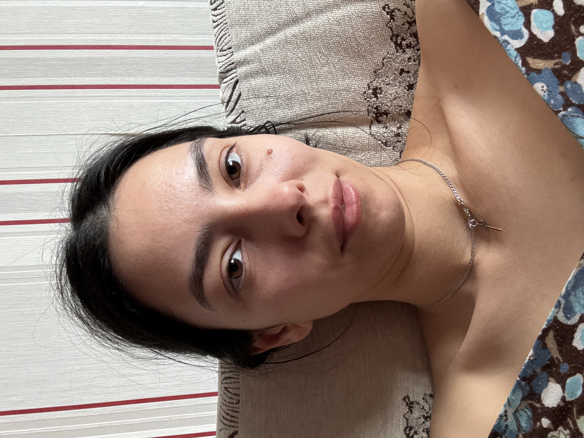A young woman is depicted in a close-up, overhead shot, lying down indoors, likely on a couch or bed. Her head is positioned to the left of the frame, and her body extends towards the right. She has dark hair, dark eyebrows, and brown eyes, looking directly at the camera with a neutral to slightly contemplative expression, a subtle hint of a smile on her lips. A small mole or beauty mark is visible on her right cheek, above her lip.
She is wearing a silver chain necklace with a small, cross-like or abstract pendant. Part of a patterned garment, featuring blue, brown, and white floral or abstract designs, is visible on her right shoulder and upper chest. She is resting on a beige, textured cushion or part of a couch, which occupies the middle and right side of the background. To the left, a wall with horizontal stripes—alternating white and thin red lines—is visible. The lighting is bright and even, suggesting daytime. No text is visible in the image.