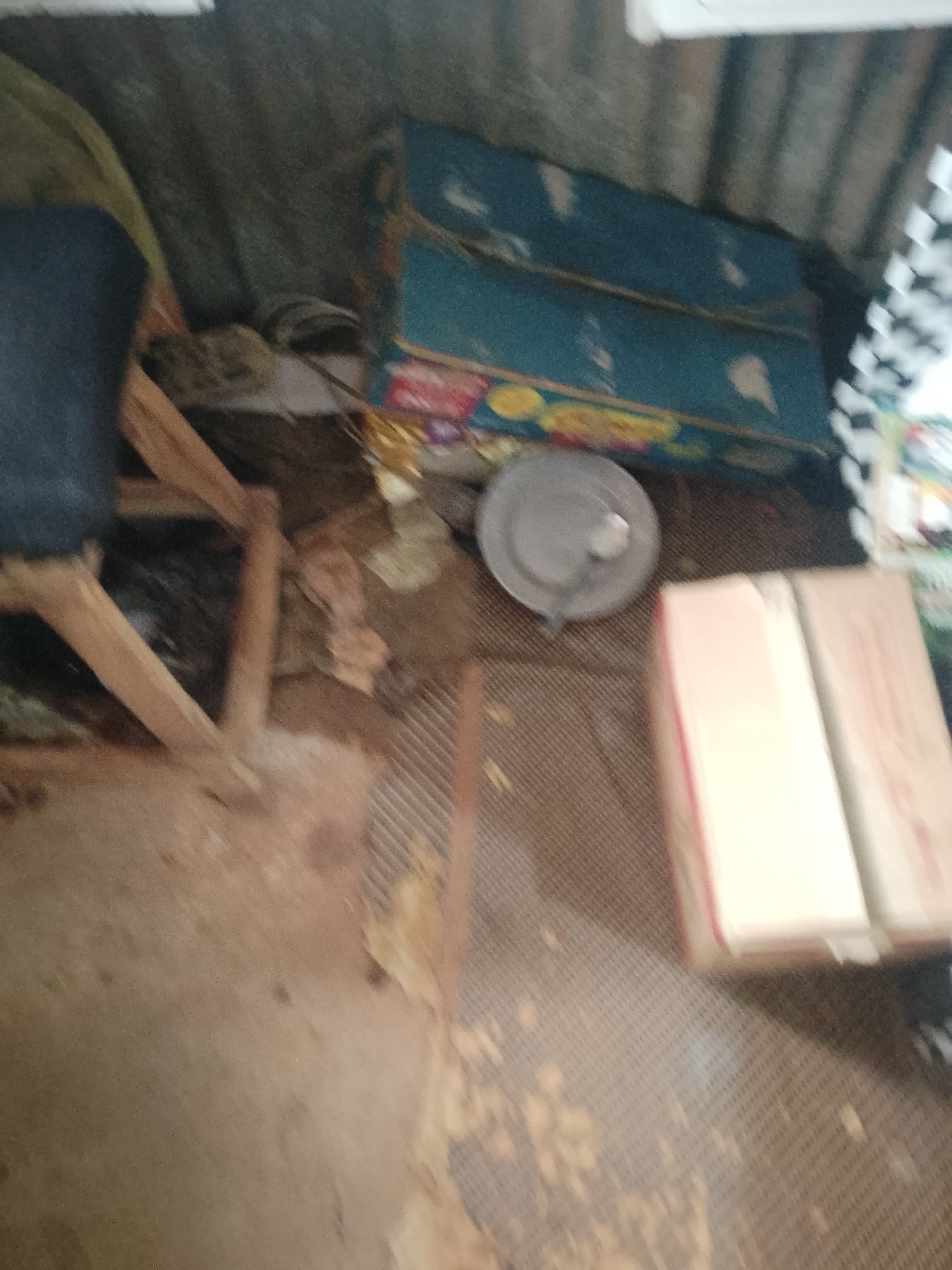 In an interior space, possibly a storage area or workshop, in Unknown, Nigeria, a scene of disarray is depicted. The ground is a mix of dirt and what appears to be wood shavings or debris, with a woven matting texture visible in some areas. To the left, a dark blue cushioned chair with wooden legs is partially visible, suggesting it has been moved or is in disuse.
Dominating the background are two large, teal-colored boxes stacked on top of each other, adorned with colorful imagery, possibly indicating they contain goods or merchandise. Next to these boxes, a metallic, round object, resembling a plate or lid, rests on the floor. Further to the right, two cardboard boxes are neatly stacked, one on top of the other, suggesting recent delivery or organization.
The walls of the space are corrugated metal, a common building material in many parts of Nigeria. The overall impression is one of a functional, yet cluttered, environment, likely a place where items are stored or processed. The lighting is dim, contributing to the somewhat hazy and indistinct quality of the image. There are no people or animals visible in the frame, and no actions or interactions are occurring. The scene is static and appears to be an unposed snapshot of the environment.