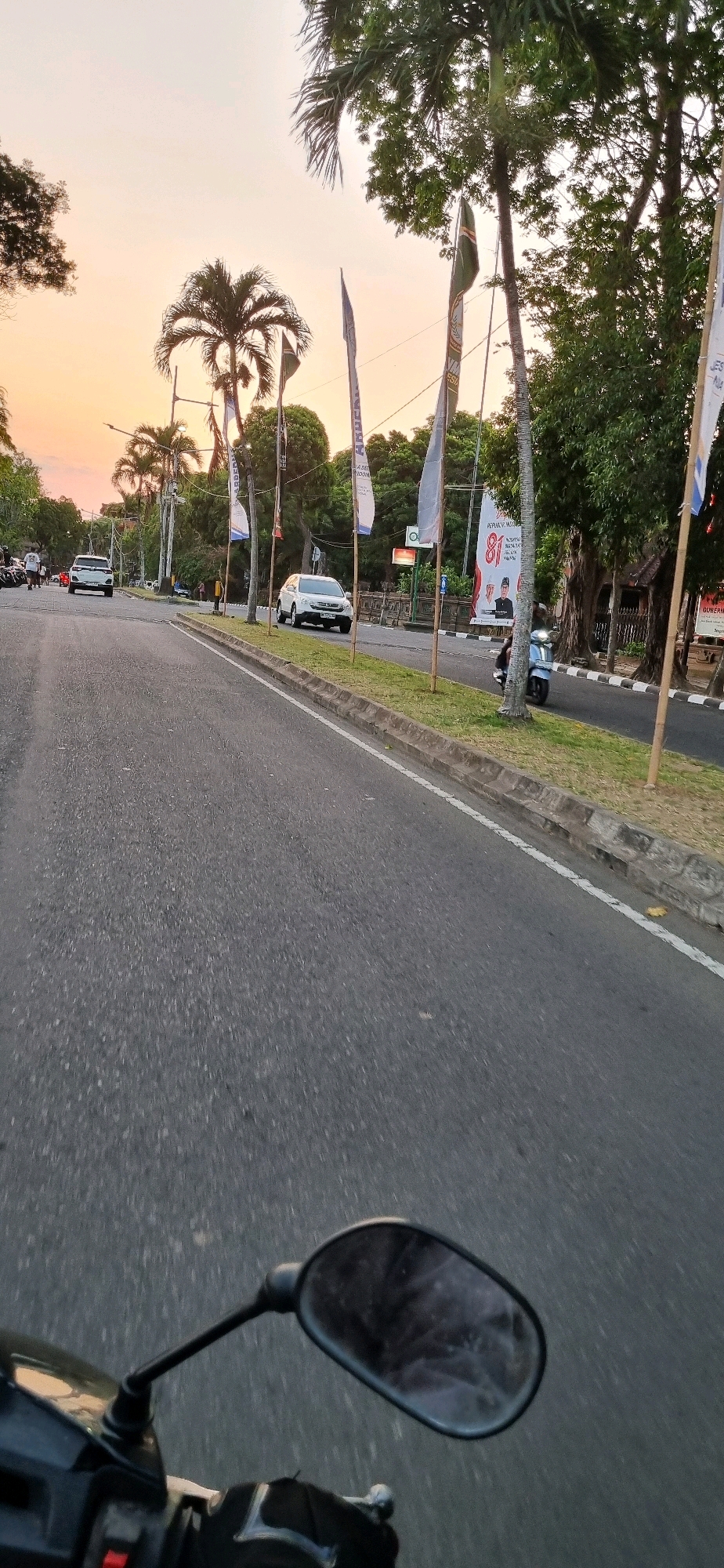 This image captures a view from a motorcycle on a road in Denpasar, Indonesia, likely during the late afternoon or early evening due to the warm, soft light and the orange hue of the sky. The road is paved and appears to be a main thoroughfare, with a white line marking the edge on the right. A grassy median separates the lanes of traffic, bordered by a concrete curb. Several palm trees and other lush green trees line the sides of the road, some with flags attached to poles in front of them.
On the road, several vehicles are visible. A white SUV is driving in the opposite lane, and further ahead, another white car is turning. A scooter is being ridden in the same direction as the camera. In the background, there are hints of buildings and more trees. A large banner with text and an image of a person is displayed prominently on the right side of the road. The foreground shows the motorcycle's mirror and handlebars, indicating the perspective of the rider. The overall atmosphere is one of everyday life in a tropical urban environment.
