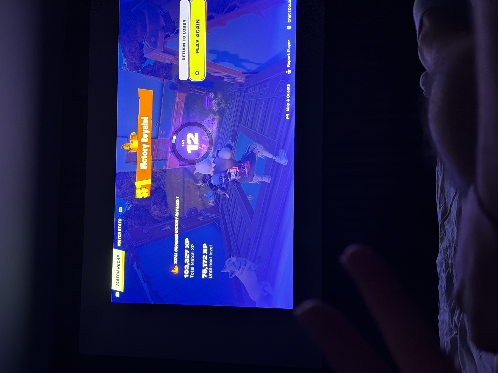 A photograph shows a television screen displaying a "Victory Royale!" screen from the video game Fortnite. The in-game character is shown in front of the "Victory Royale!" message, along with information about total match XP (102,327 XP) and XP until the next level (75,172 XP). Options to return to the lobby or play again are visible, as are the player's current level, level 12, and icons for map and quests, reporting a player, and chat. The environment is a dark room and a person’s hand is visible in the foreground, indicating that the picture was taken by a person. There is no indication of Pernik, Bulgaria, in the image.