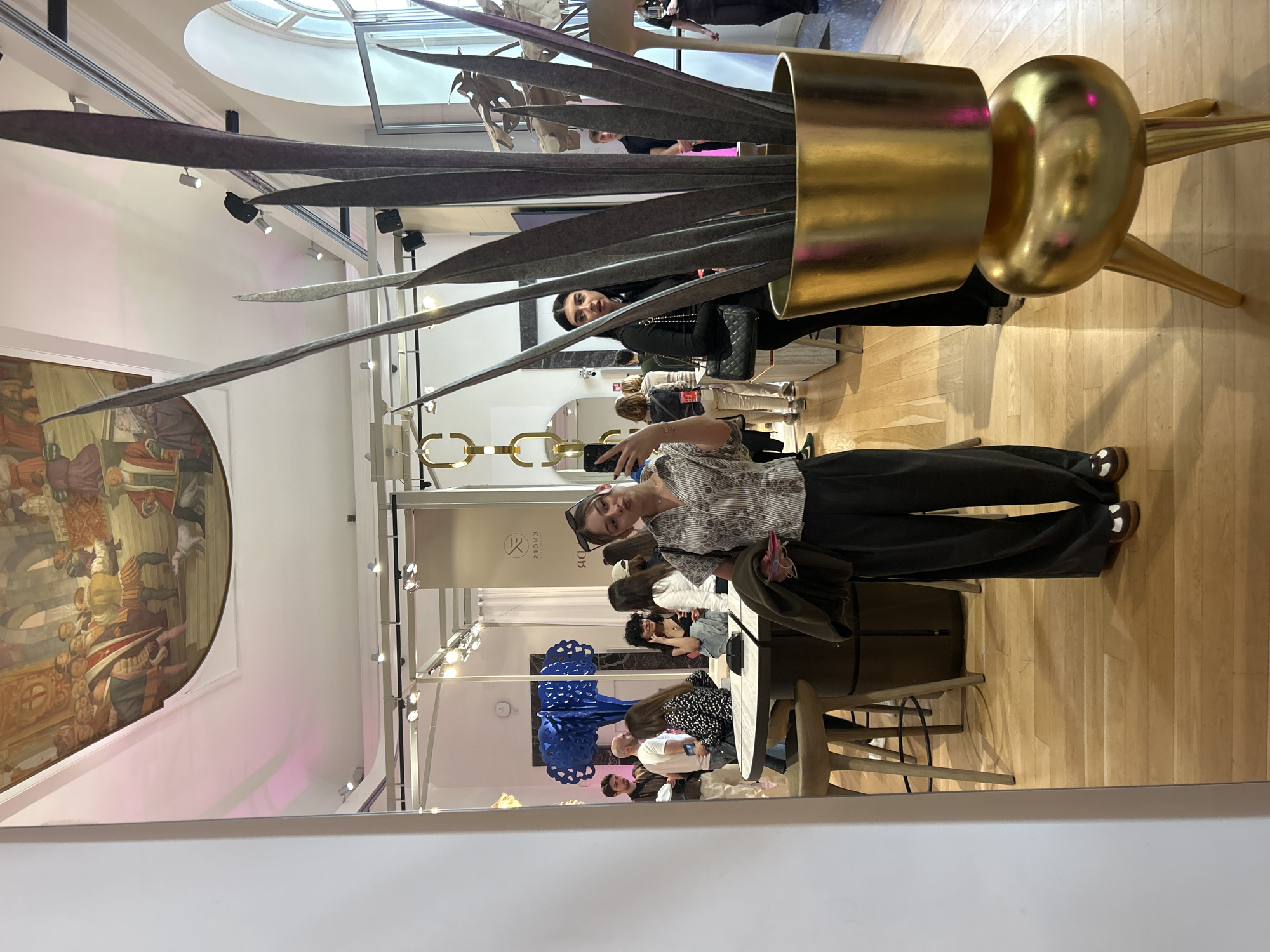 The image, taken in an indoor setting in Milan, Italy, captures an event or exhibition space with several attendees and prominent decorative elements. The photo appears rotated, showing a perspective from above looking down into the room.
In the foreground, a person is taking a selfie, reflected in a large mirror. They are wearing a light-colored striped shirt, dark trousers, and brown and white loafers, with sunglasses resting on their head. Directly behind them, partially obscured by a large sculpture, another individual with dark hair is visible, wearing a dark top and carrying a quilted black handbag.
The central focal point is a large, abstract sculpture or art installation. It consists of a shiny golden cylindrical base that transitions into a golden sphere on one side. Extending from this base are numerous long, slender, dark grey, felt-like blades or leaves that stretch upwards and outwards.
In the background, several other people are seated at tables, some appearing to be engaged in conversation, while others look at their phones. The overall atmosphere seems relaxed and social. The venue features light wooden flooring, white walls, and a high ceiling with a large, circular skylight, suggesting it is daytime. A large, colorful mural depicting a historical or classical scene with multiple figures is visible on one of the walls. Modern spotlights are installed along the ceiling.
On a white sign reflected in the mirror, partially obscured and inverted text is visible, possibly reading "KIOSK" and "DB" with an accompanying logo, likely indicating a brand or event organizer. The stylish decor and gathering of people suggest a fashion, design, or art event.