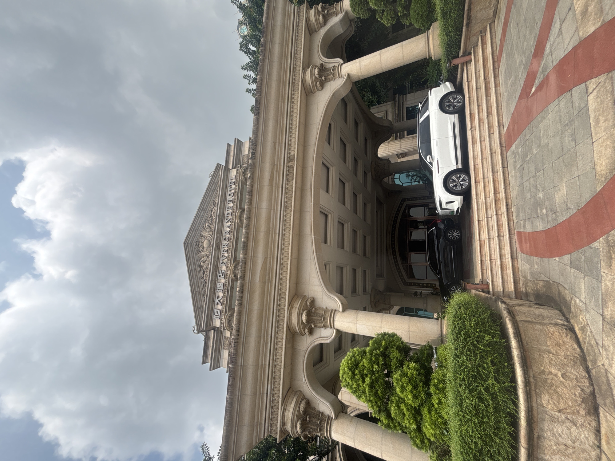 The image shows the grand entrance of the Evergrande Hotel in Nanhai District, China. A large, ornate building with classical architecture, featuring massive columns and intricate carvings, is visible. Two luxury vehicles, a black SUV and a white SUV, are parked on the steps leading up to the entrance. Lush green bushes and trees surround the entrance, with a stone planter in the foreground. The sky is overcast with large clouds. The overall scene suggests opulence and a formal atmosphere.