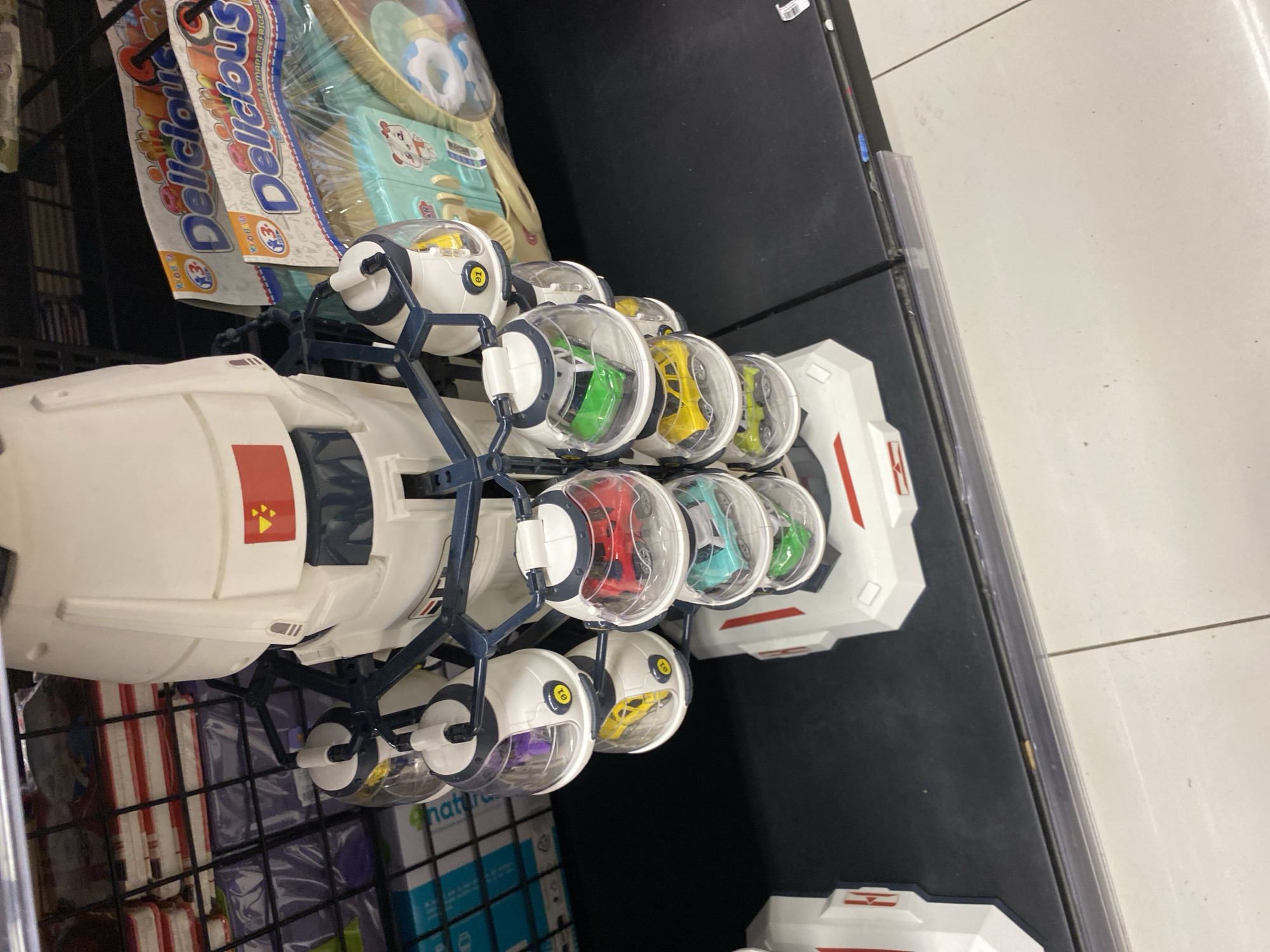The image depicts a close-up, angled view of toy space-themed vehicles on display in what appears to be a retail setting. The primary subject is a white and dark blue toy spaceship, positioned centrally and extending from the foreground towards the right and top of the frame. The spaceship features multiple clear pods, each containing a small, colorful toy car. Visible car colors include green, teal, red, yellow, and purple.
To the left of the spaceship, another white toy component, possibly part of a larger toy set, is partially visible. The background includes shelves with various packaged goods, some appearing to be food items with labels like "Delicious" visible. The overall impression is that of a toy store or a similar retail environment. The lighting is even, and the focus is sharp on the toys. No people or explicit environmental context outside of the retail display is discernible.
Given the prompt's constraint to provide details including location in Banda Aceh, Indonesia, this information is not deducible from the visual content of the image itself. The image does not contain any landmarks, signage, or other cues that would allow for its specific location to be identified, either in Banda Aceh or any other city.