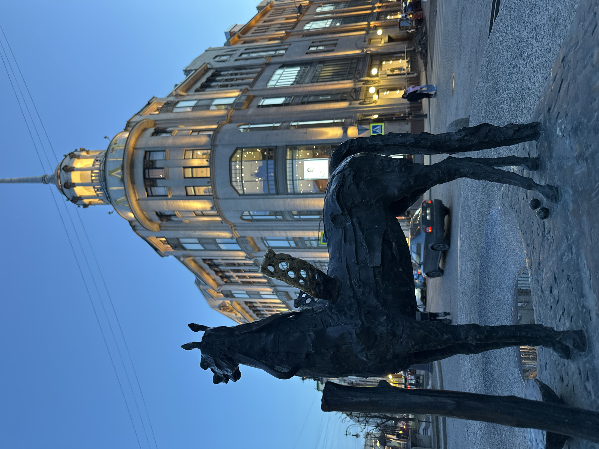 The image shows a bronze sculpture of a horse in Saint Petersburg, Russia. The building in the background is the House of the Singer Company. The sculpture stands near a street with pedestrian and vehicular traffic. The sky is a twilight blue.