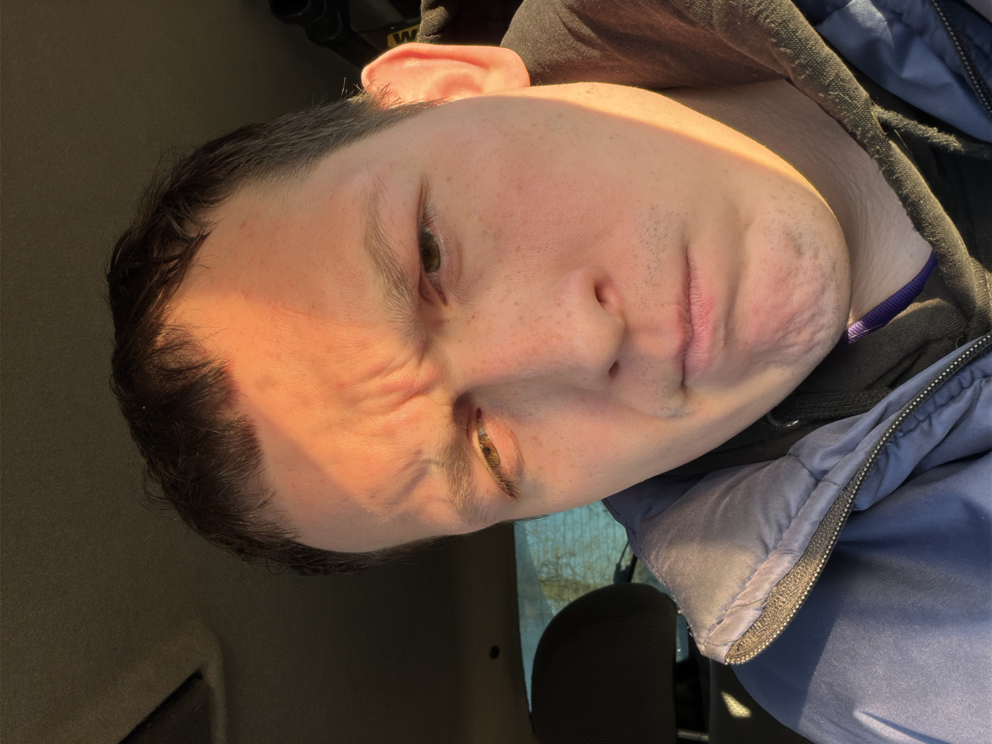 A man is indoors in Chaikovsky, Russia. He is wearing a dark hoodie and a blue puffer jacket. His face is illuminated by sunlight. The background shows a partial view of a car interior.