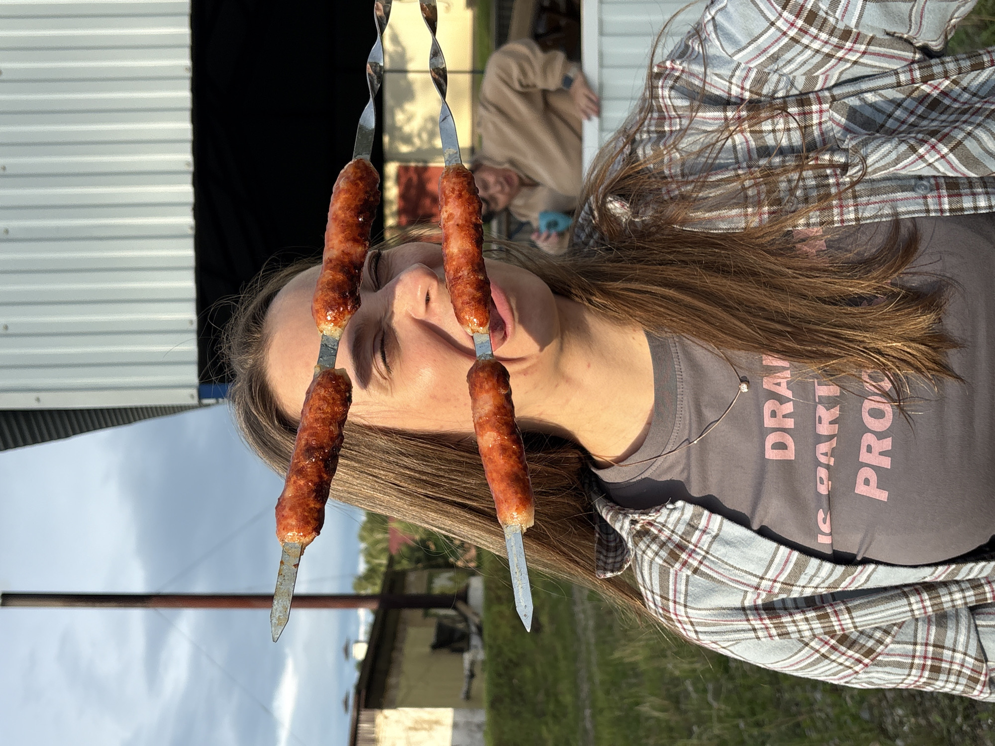 A young woman with long brown hair is pictured outdoors, appearing to be in Birobidzhan, Russia, based on contextual clues. She is wearing a grey t-shirt with pink lettering that reads "DRAIN IS PART OF PRO" and a plaid shirt open over it. Her face is contorted as if she is about to eat sausages on skewers, with three skewers positioned horizontally in front of her face. One sausage is near her eyes, another is by her mouth, and a third is lower down. In the background, another person in a tan hoodie is visible, and a building with a corrugated metal roof is present, along with some greenery and a cloudy sky. The overall scene suggests a casual, outdoor gathering.