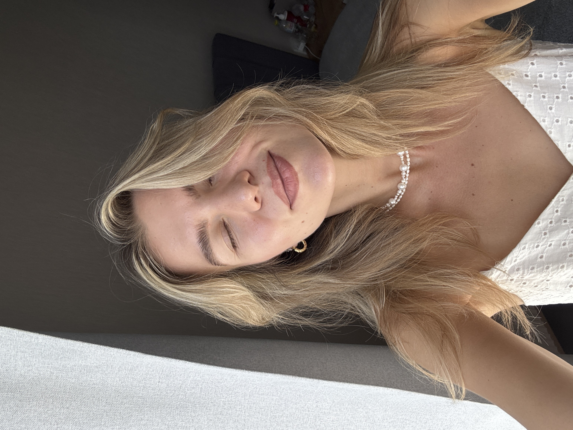 An overhead, medium shot shows a young woman with blonde hair, her eyes closed and a slight smile on her face, illuminated by natural light. She wears a white, eyelet camisole and a double-strand pearl necklace. To the left of her face, a textured grey curtain casts a shadow. The background is dark, with the corner of a bed and a nightstand visible on the right. The image is taken in an indoor setting, likely a bedroom.