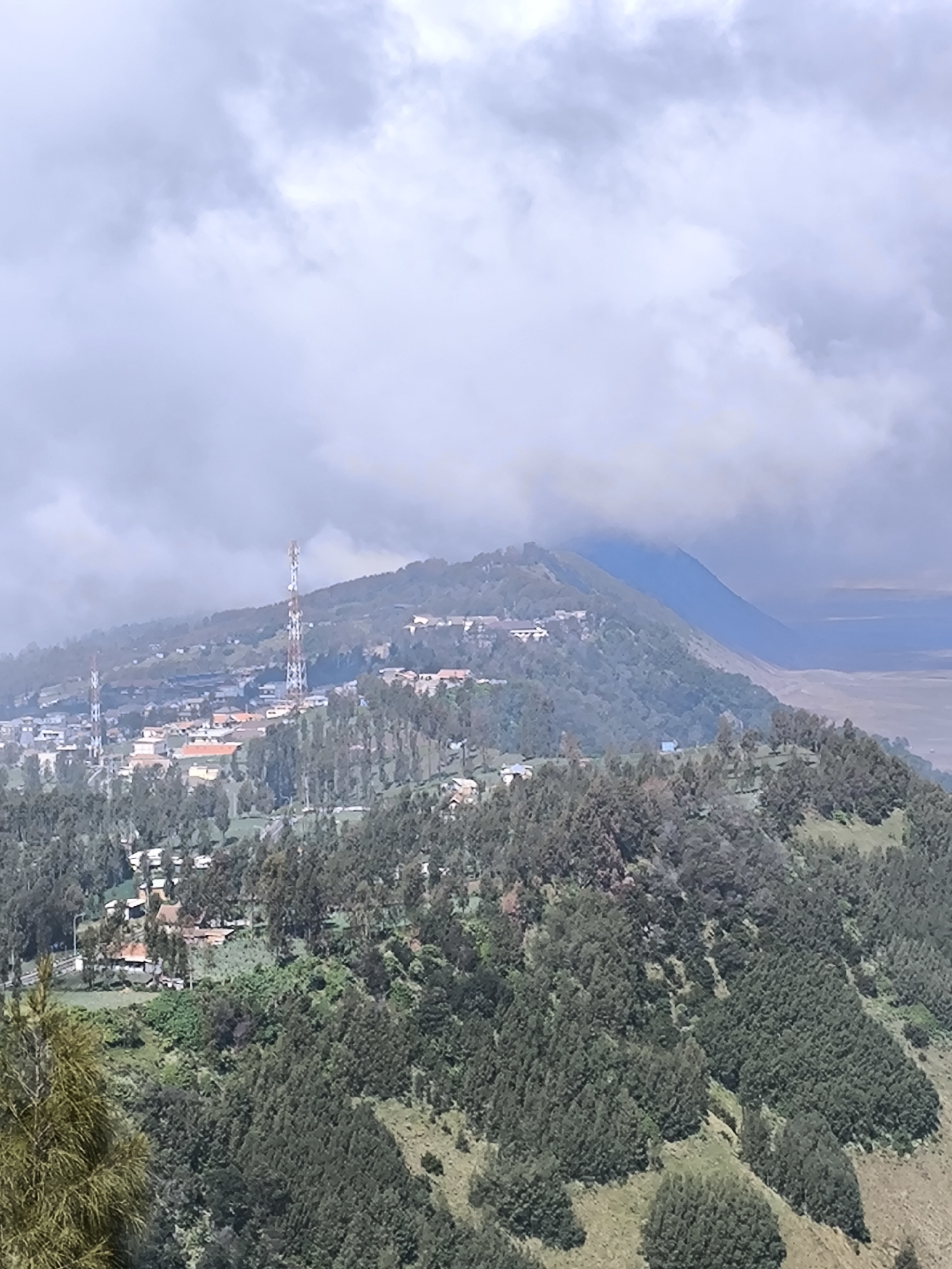 The image depicts a scenic view of a village nestled on a lush green mountainside in Cemorolawang, Indonesia. The foreground is dominated by dense green trees, with some lighter green grasses visible on the slopes. In the midground, a village is spread across the hillside, characterized by numerous houses with terracotta-colored roofs and some taller buildings. Two prominent telecommunication towers, one taller and more detailed than the other, stand out against the backdrop of the village and the mountains. The village appears to be a residential and possibly commercial area, with a road visible leading into it.
Behind the village, rolling mountains covered in vegetation extend into the distance. The highest peak in the background is partially obscured by dramatic, thick clouds, suggesting cloudy or foggy weather. The clouds are a mix of light and dark grays, creating a dynamic sky. The overall impression is one of a tranquil, high-altitude settlement amidst a vast natural landscape. The lighting suggests it is daytime, with diffused light indicating potential overcast conditions or early morning/late afternoon sun. There is no visible text in the image. The scene evokes a sense of peace and natural beauty, with the human settlement integrated into the mountainous terrain.