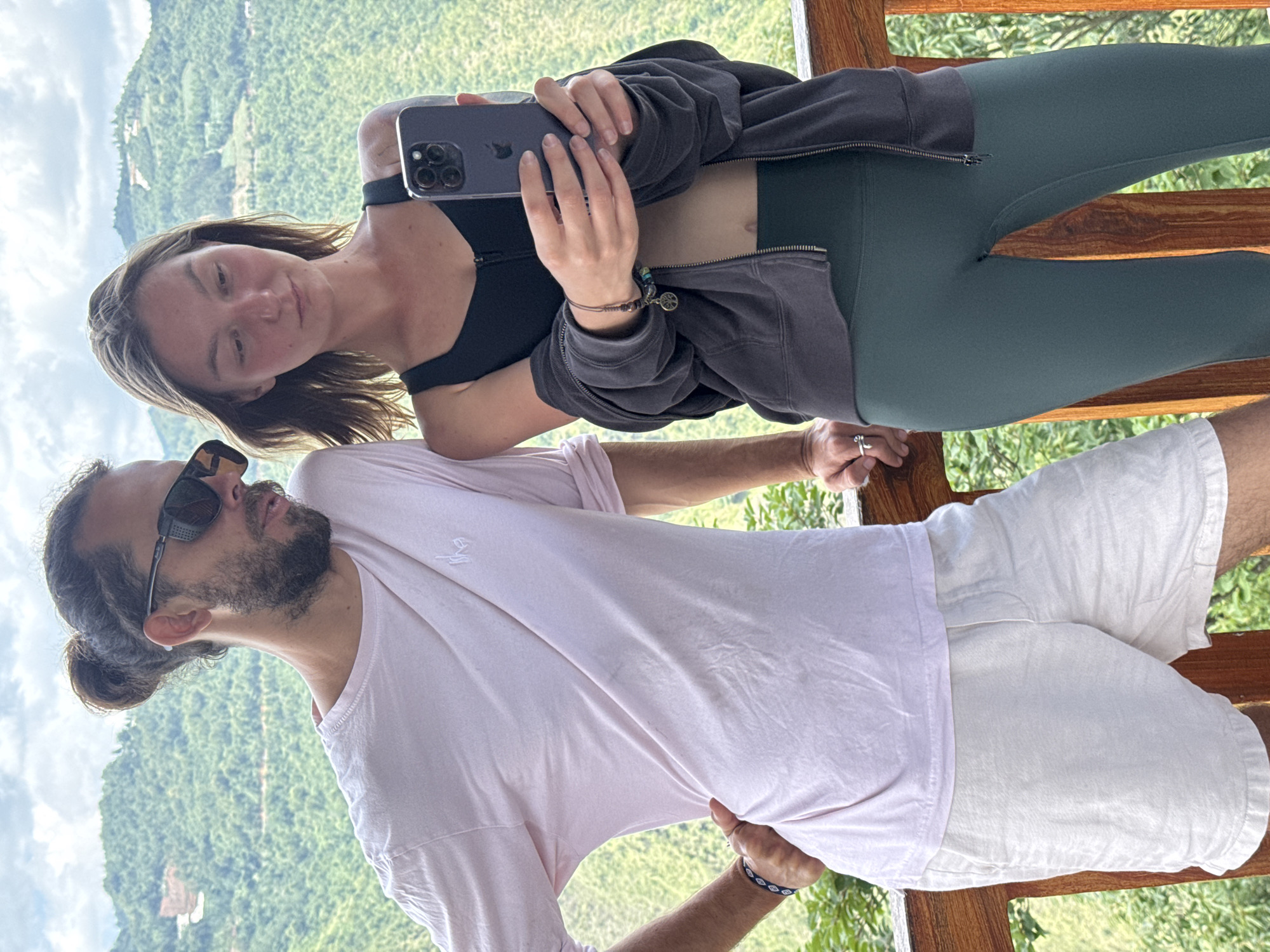 Two people stand on a wooden balcony overlooking a lush green landscape. A woman holds a smartphone, possibly taking a selfie, and leans against a man. The woman wears a black sports bra, a dark jacket, and dark green leggings. The man wears sunglasses, a pink shirt, and white shorts. The background reveals a green, mountainous terrain with hints of buildings, suggesting a location in or near a populated area. The scene is set during the daytime with natural light. The location is likely Lũng Cú Commune, Vietnam, based on the geographical description and identifying cultural characteristics.