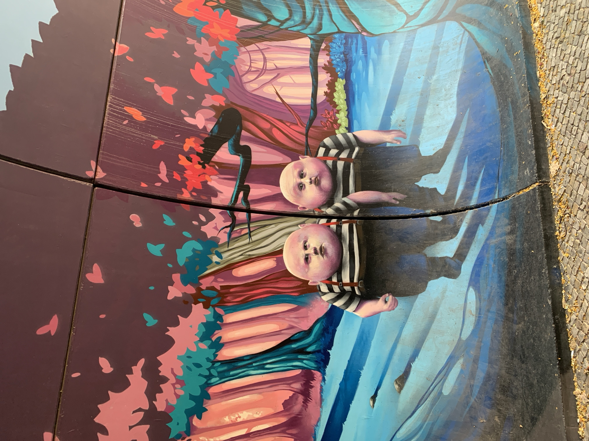 This is an outdoor mural painted on a curved surface, possibly a bench or a decorative element, set against a cobblestone ground. The mural depicts a surreal, dreamlike scene with two pale-skinned, bald figures reminiscent of infants, wearing striped shirts and suspenders. Their faces have a slightly bewildered or unnerving expression. One figure lies on its back with arms outstretched, while the other is positioned similarly, with only its head and torso visible.
The background is a vibrant and abstract landscape. To the left, there's a dark, stylized tree with red and pink leaves, set against a purple backdrop. To the right, a blue, watery landscape with abstract shapes and reflections is depicted. The overall style is painterly and stylized, with bold colors and fluid lines.
The image is taken from a slightly elevated angle, and the mural is divided into sections by visible seams or cracks. The lighting suggests it is daytime, with no indication of specific weather conditions beyond what the natural sunlight illuminates. There are no visible text elements in the mural or its surroundings. The setting appears to be a public space, given the cobblestone pathway. The surreal and unsettling depiction of the figures, combined with the fantastical landscape, creates a mysterious and perhaps slightly disquieting atmosphere.