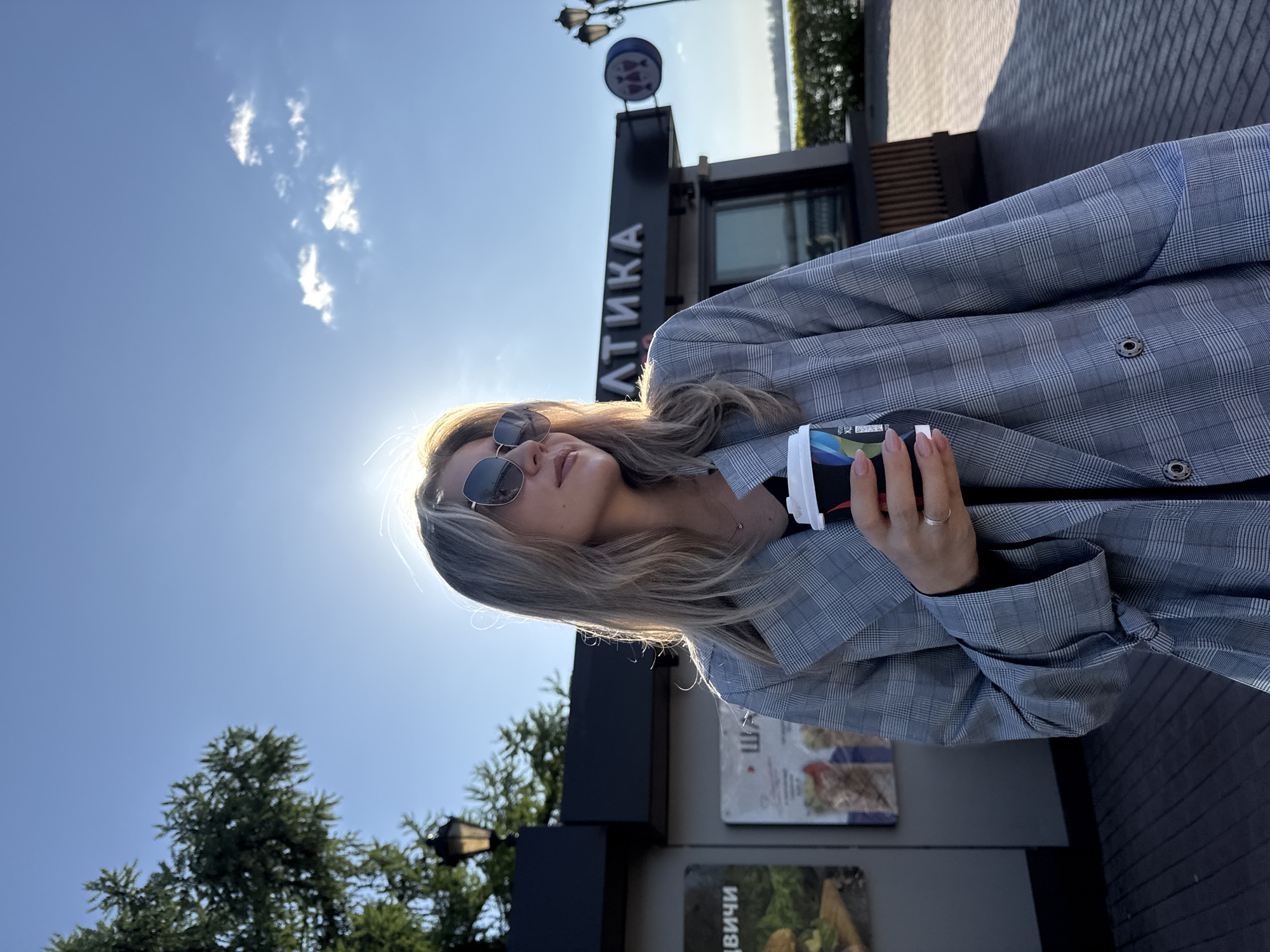 A woman is standing outdoors in Unknown, Unknown, holding a cup. She is wearing sunglasses and a grey plaid jacket. The sun is shining brightly behind her. There is a building with the word "ЛТИКА" in Cyrillic on it, and a lamp post nearby. Trees and other vegetation are also visible.