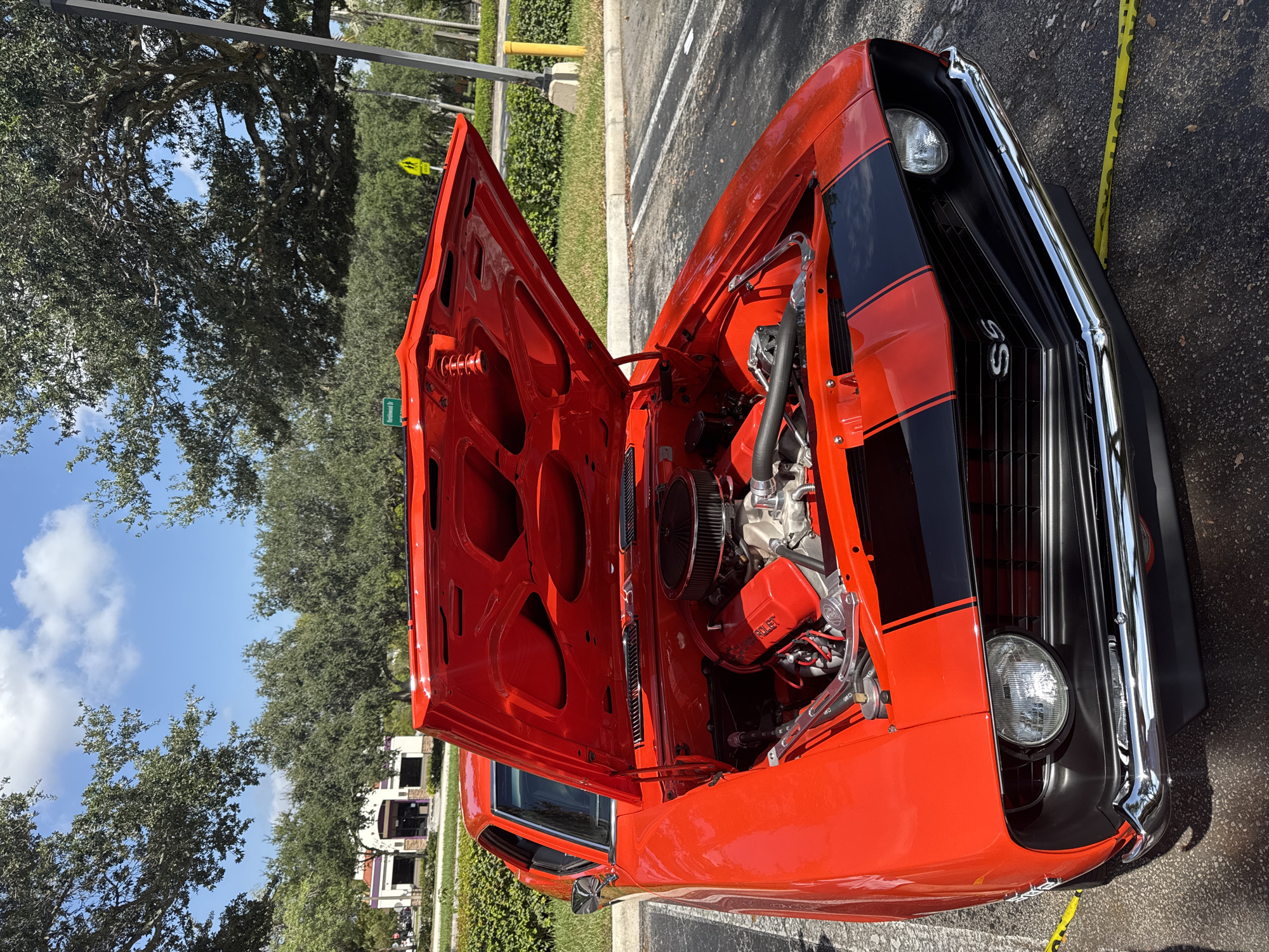 A vibrant red 1969 Chevrolet Camaro SS is parked in a paved lot under a clear blue sky with scattered clouds. The car's hood is open, revealing a powerful engine with chrome accents and a black air filter. Black racing stripes adorn the hood. The car is positioned at an angle, with its front end facing the viewer. In the background, trees line the edge of the lot, and a building with a purple awning is visible. Yellow caution tape is laid out on the ground in front of the car. The location is the parking lot of the Bank of America building at 9000 W Sample Rd, Coral Springs, Florida, United States of America.