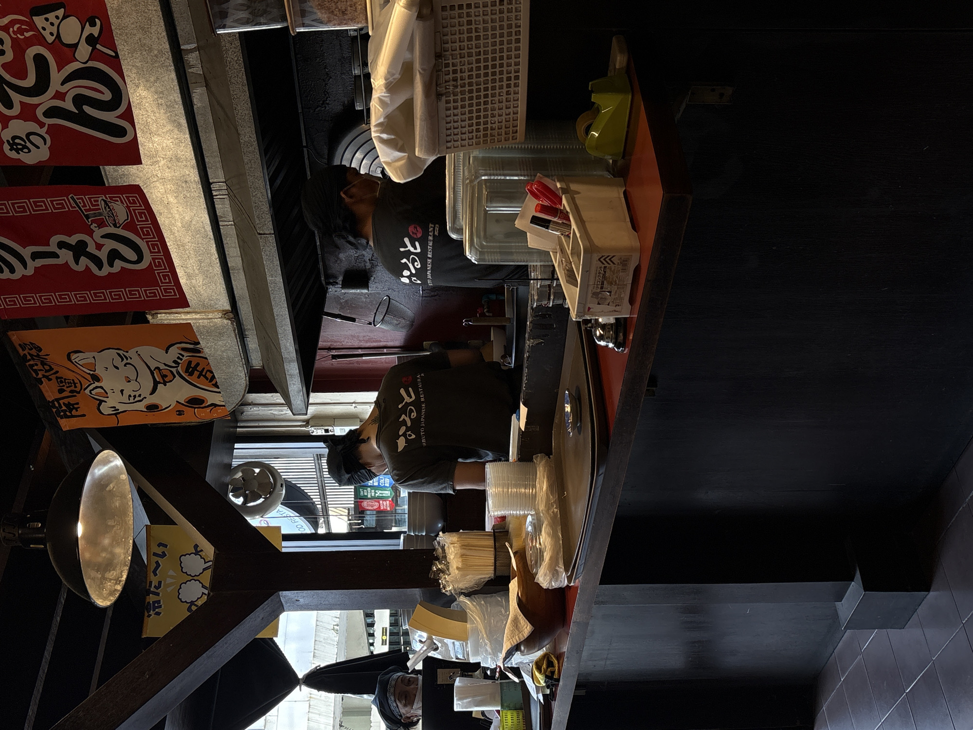 The image depicts the interior of a Japanese restaurant, likely in Chiang Mai, Thailand. The scene is dimly lit, suggesting it could be evening or indoors with artificial lighting. A counter area is visible, with a person wearing a dark uniform and a black bandana or cap serving. They appear to be working behind the counter, possibly preparing food or taking orders, though their face is not fully visible.
Various items are present on the counter, including stacks of plastic cups, chopsticks in wrappers, and condiments. To the left, several red banners with Japanese characters are hanging, some featuring illustrations of food items. An orange banner with a "maneki-neko" (beckoning cat) illustration is also visible. Above the counter, a large, metallic lamp hangs down, illuminating the area. In the background, what appears to be a kitchen or service area is visible, with shelves and equipment. Another person, partially visible at the bottom, is also wearing a black head covering. The overall atmosphere seems to be that of a bustling, functional restaurant environment. Text in Japanese characters is visible on the banners, likely indicating menu items or the restaurant's name. The text "ARUTO JAPANESE RESTAURANT" is also visible on the uniform.